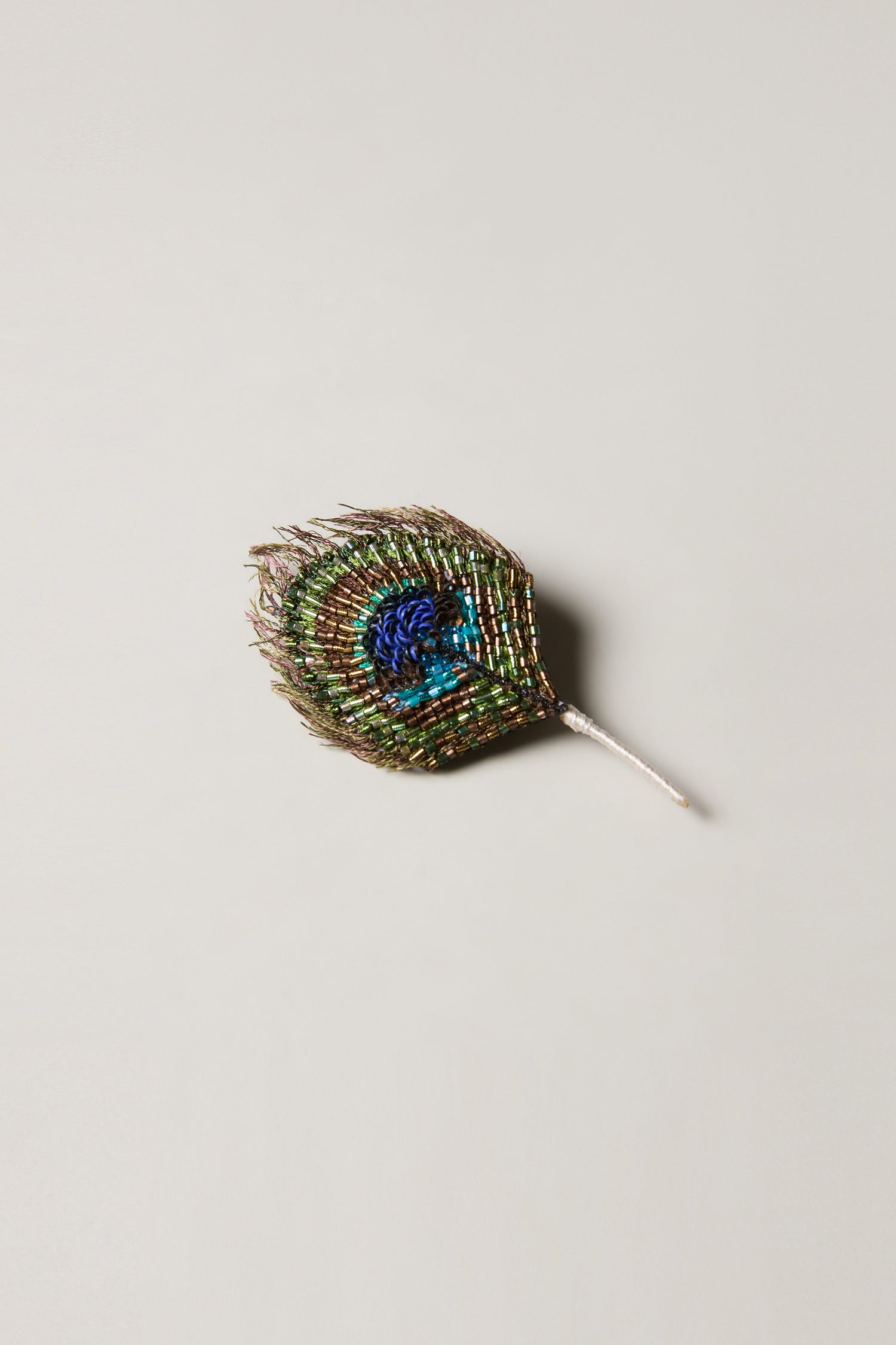 The Hand Beaded Peacock Feather Brooch, featuring iridescent green, blue, and gold tones, is showcased on a simple light background.