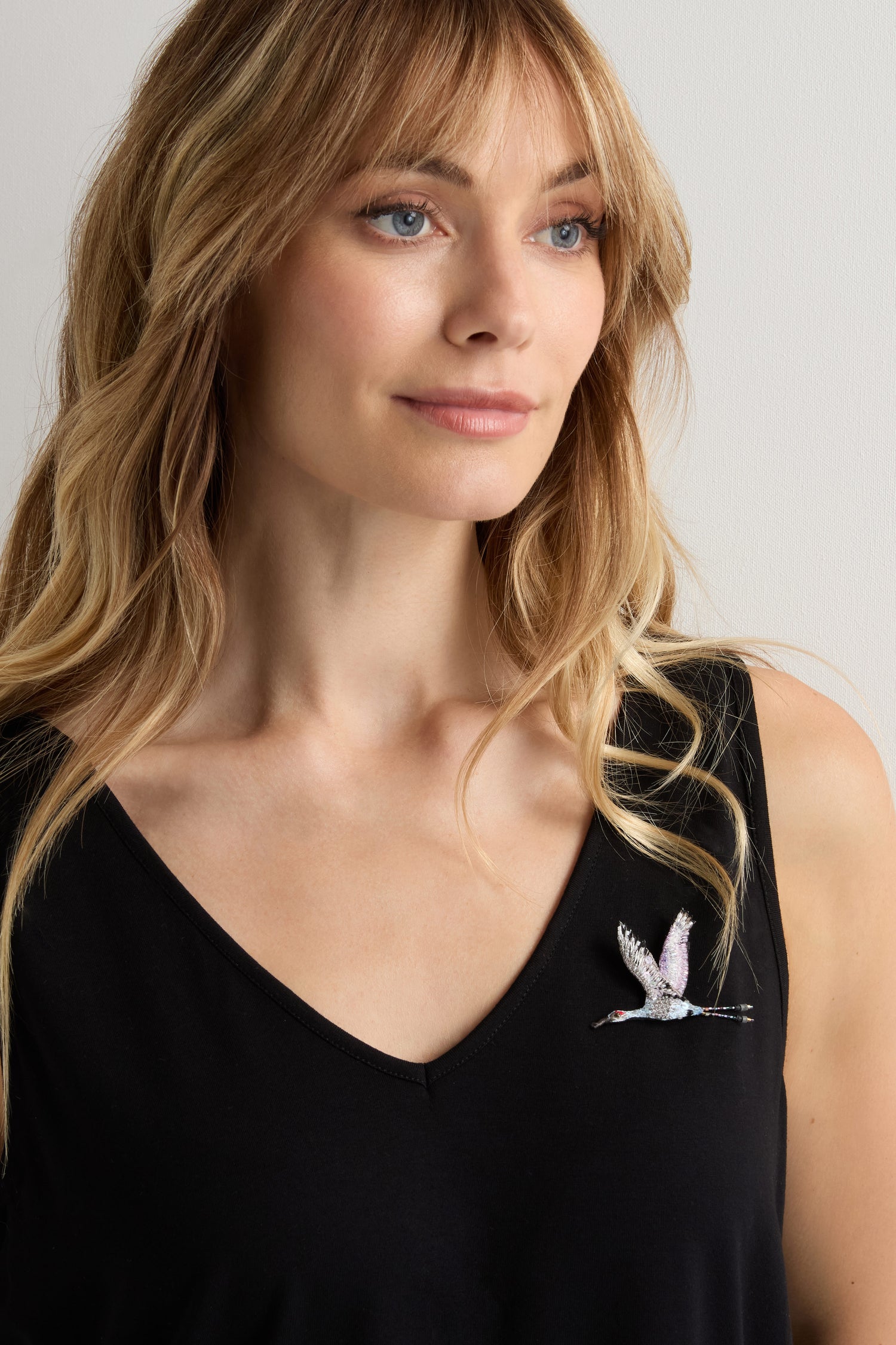A woman with long blonde hair wears a black V-neck top styled with the Hand Beaded Sandhill Crane Brooch, featuring a nature-inspired flying bird design.