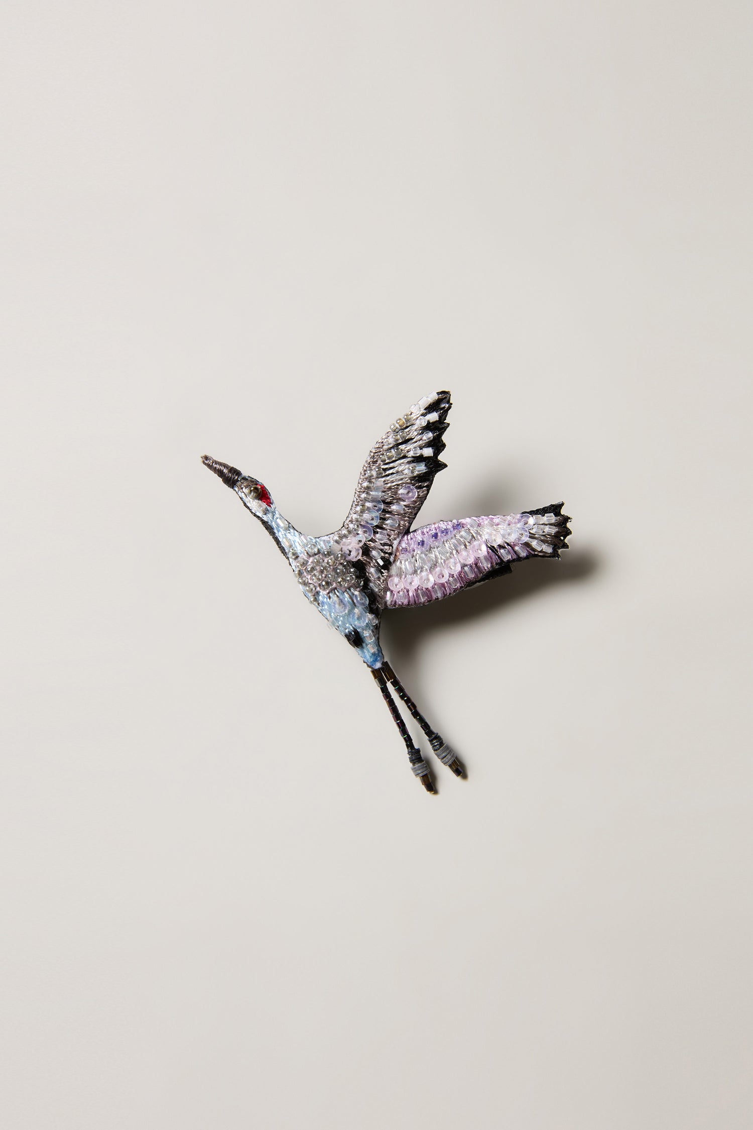The Hand Beaded Sandhill Crane Brooch features a nature-inspired crane with outstretched wings, detailed in white, pink, blue, and black beads on a light background.