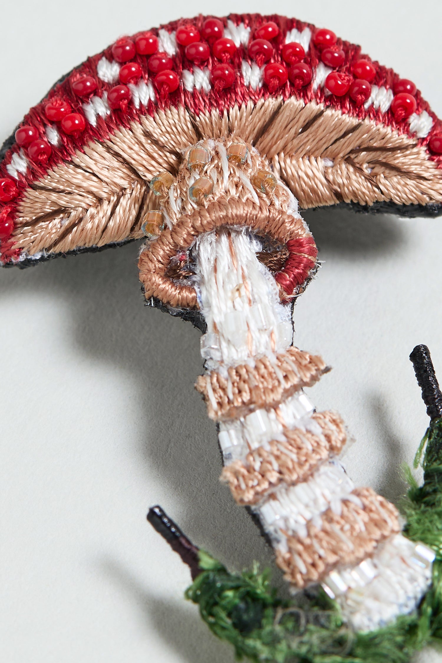 The Hand Embroidered Fly Mushroom Brooch showcases a red and white mushroom with textured stitching and delicate bead details—a striking accessory for any collection.