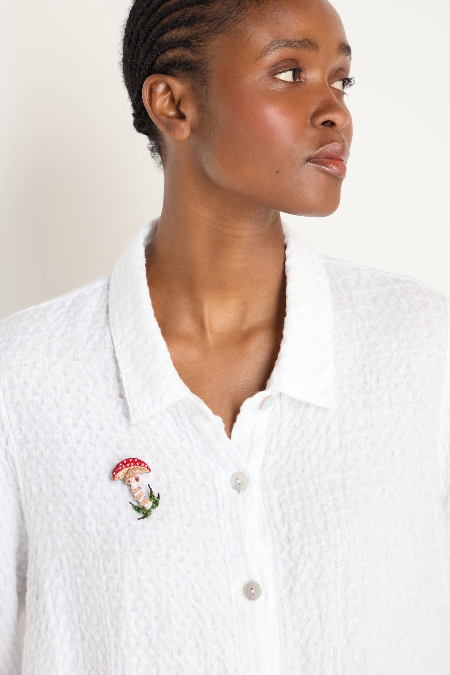 A person in a textured white button-up shirt wears the Hand Embroidered Fly Mushroom Brooch pinned on the left side of their chest.