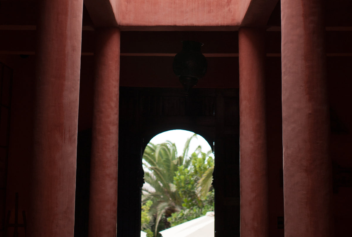 Dimly lit interior with tall terracotta columns framing an arched doorway, revealing a glimpse of green palm trees outside
