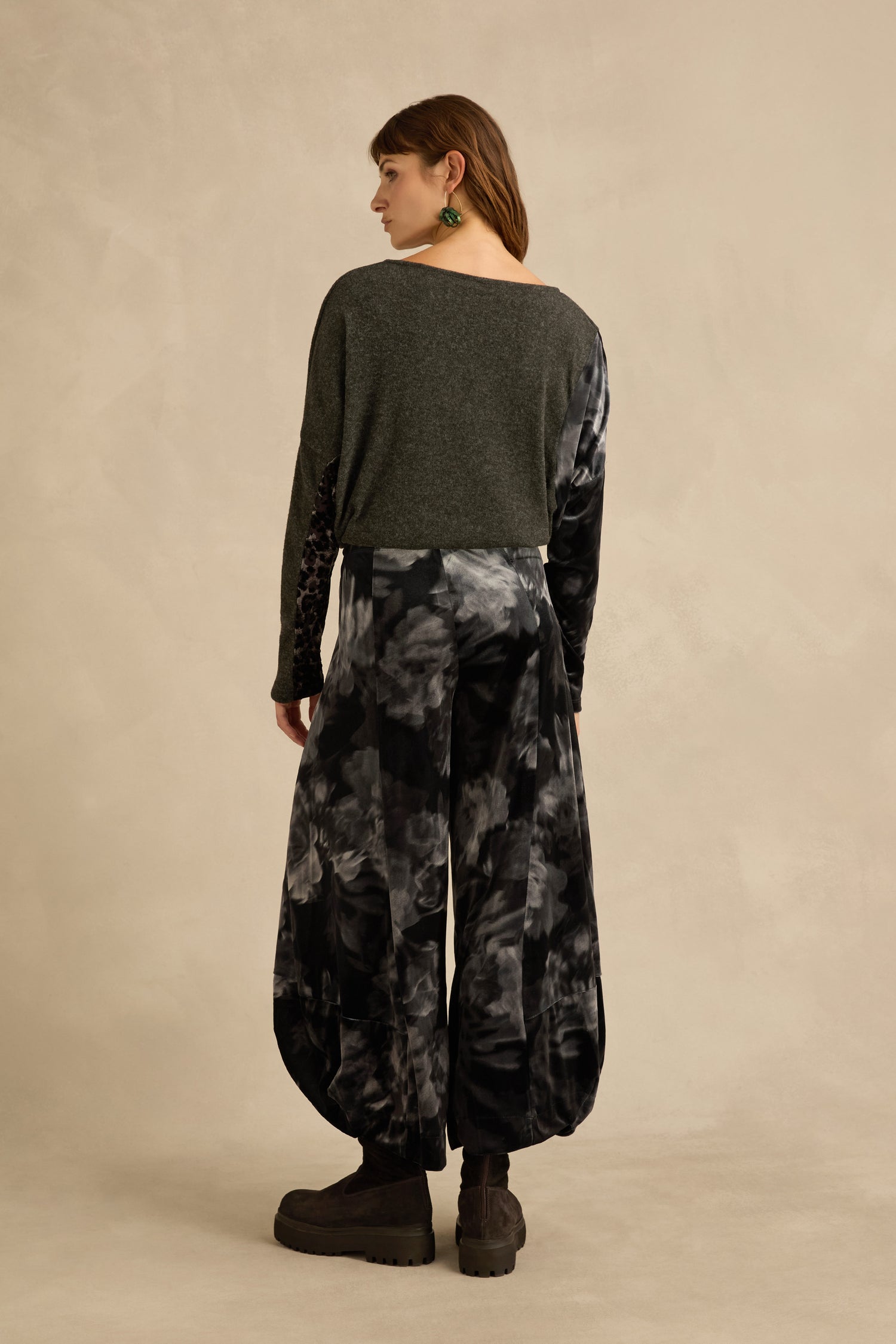 A person stands with their back to the camera, wearing a dark gray long-sleeve top, Blurred Floral Velvet Bubble Trousers in black-and-gray tie-dye with sculptural volume, platform boots, and green earrings against a beige background.