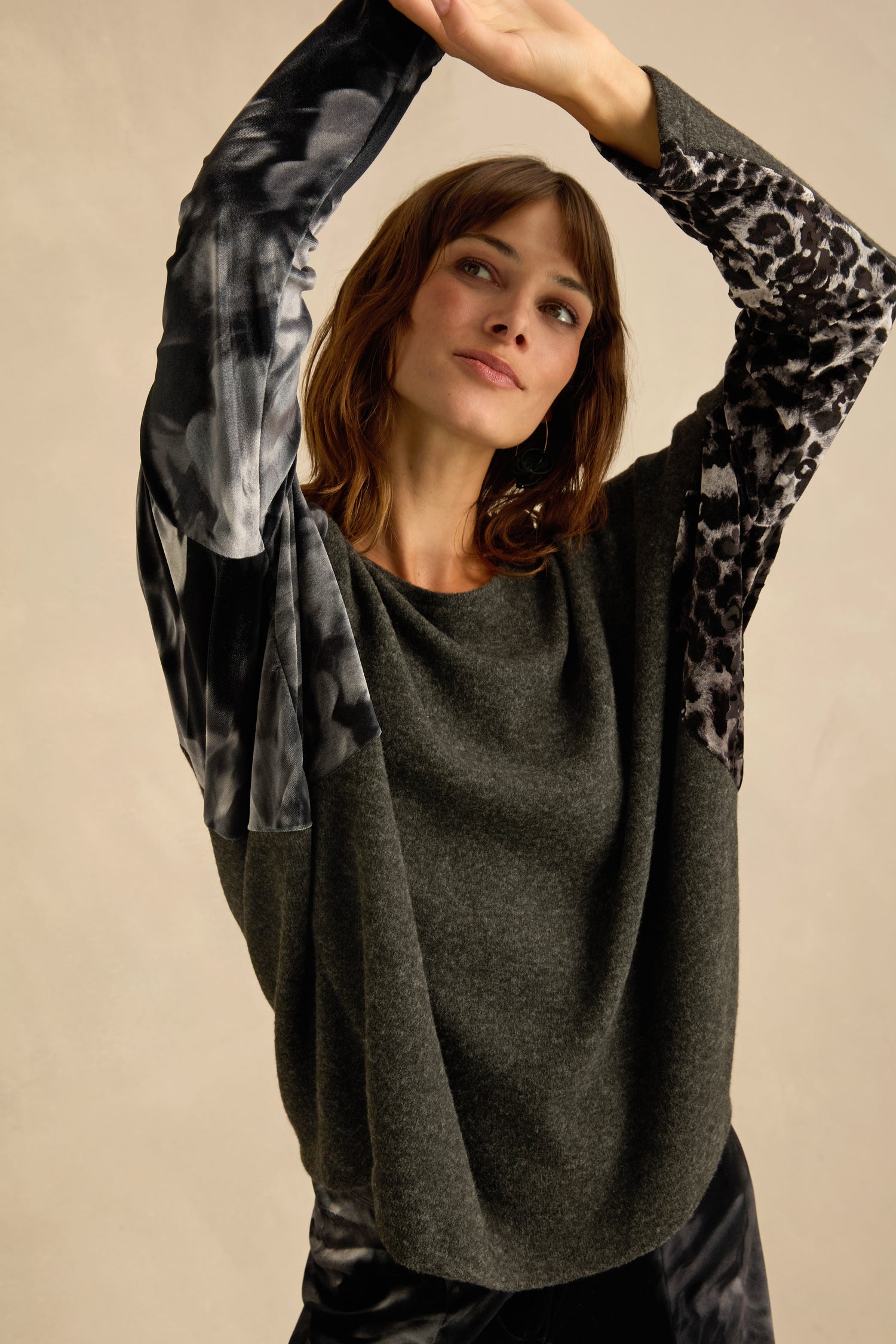 A woman poses with arms raised, wearing the Blurred Floral Jersey Top—a charcoal grey sweater featuring printed black and white sleeves—standing against a plain beige background.