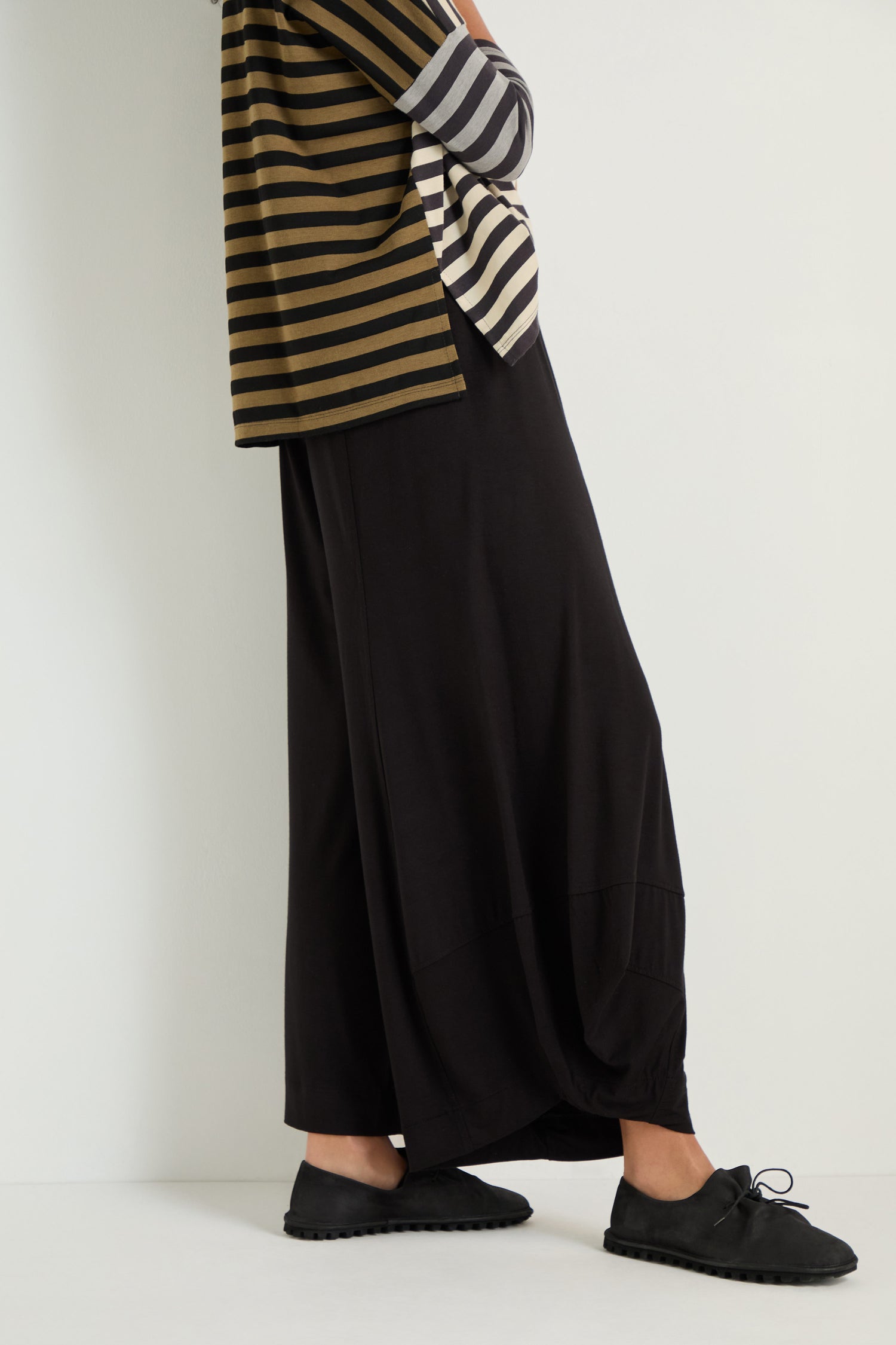 Someone wearing Bubble Hem Trousers, a striped long-sleeve top, and black lace-up shoes stands against a plain white wall.