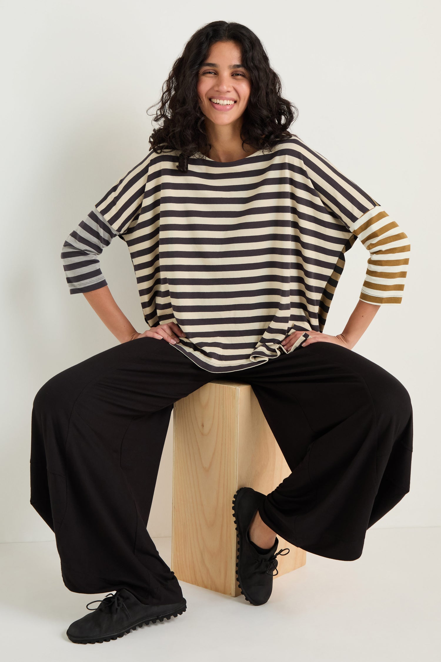 A woman smiles while sitting on a wooden block, wearing relaxed fit black Bubble Hem Trousers and a loose striped top, with her hands resting on her knees.