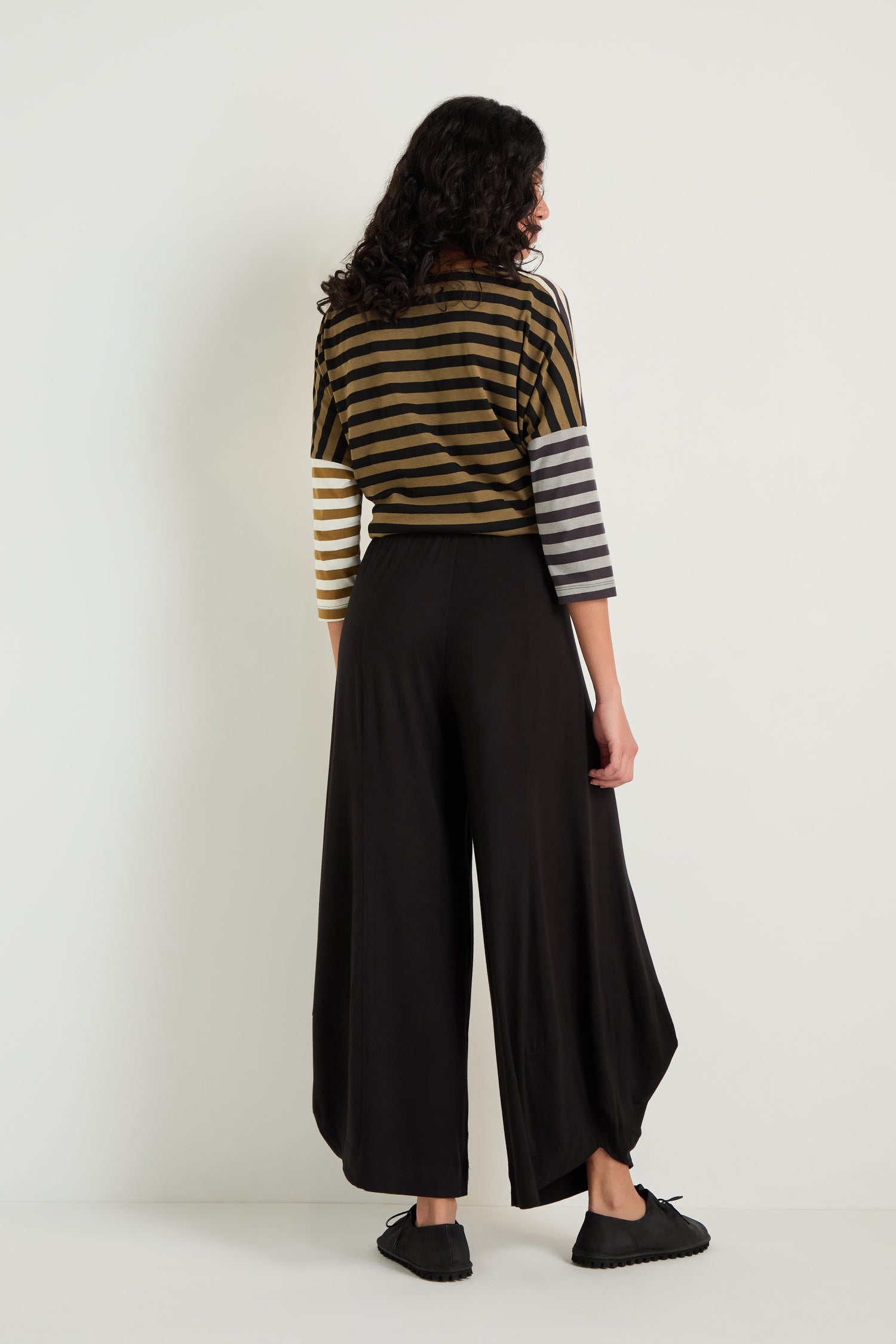 A person with long, curly hair faces away, wearing Bubble Hem Trousers and a striped stretch jersey top with three-quarter sleeves, set against a plain light background.