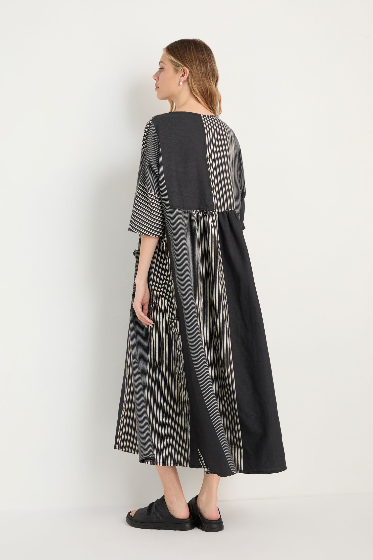 Wearing the Mixed Stripe Easy Dress—a long, loose-fitting black and white striped piece with wide sleeves—and black slip-on shoes, a person stands against a plain light-colored background.