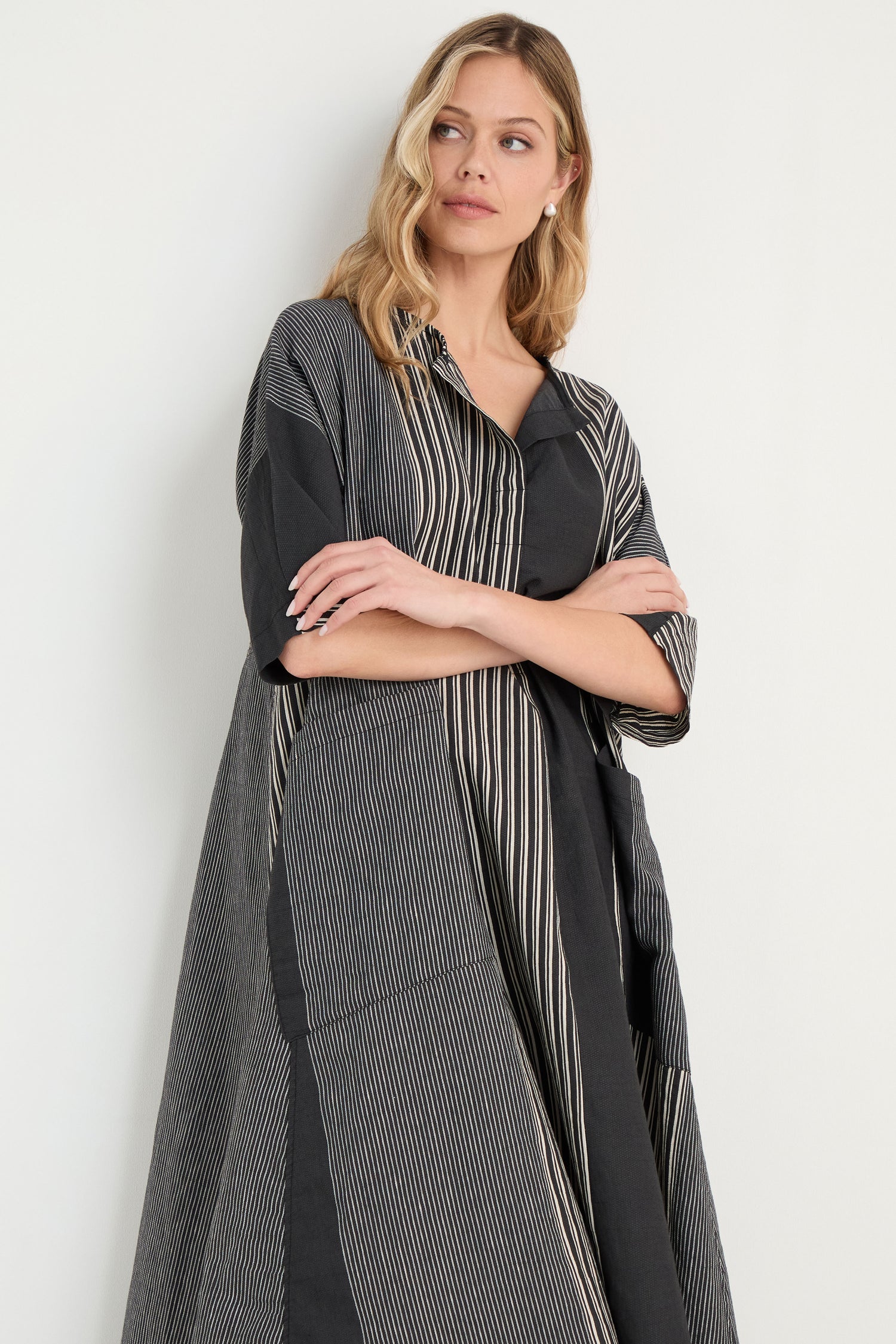 A woman stands against a light background, smiling and looking to the side in the Mixed Stripe Easy Dress—a relaxed dress featuring mixed stripes, front pockets, and elbow-length sleeves.