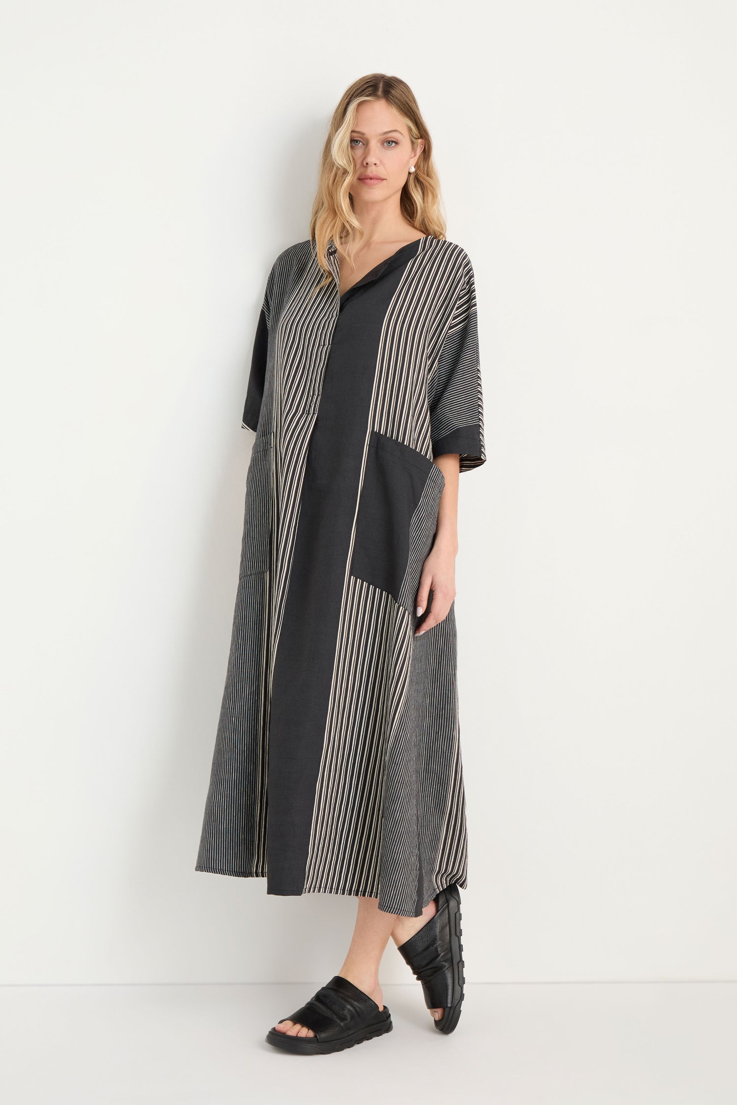A woman stands sideways, wearing the Mixed Stripe Easy Dress with vertical stripes and three-quarter sleeves, paired with black slip-on shoes.