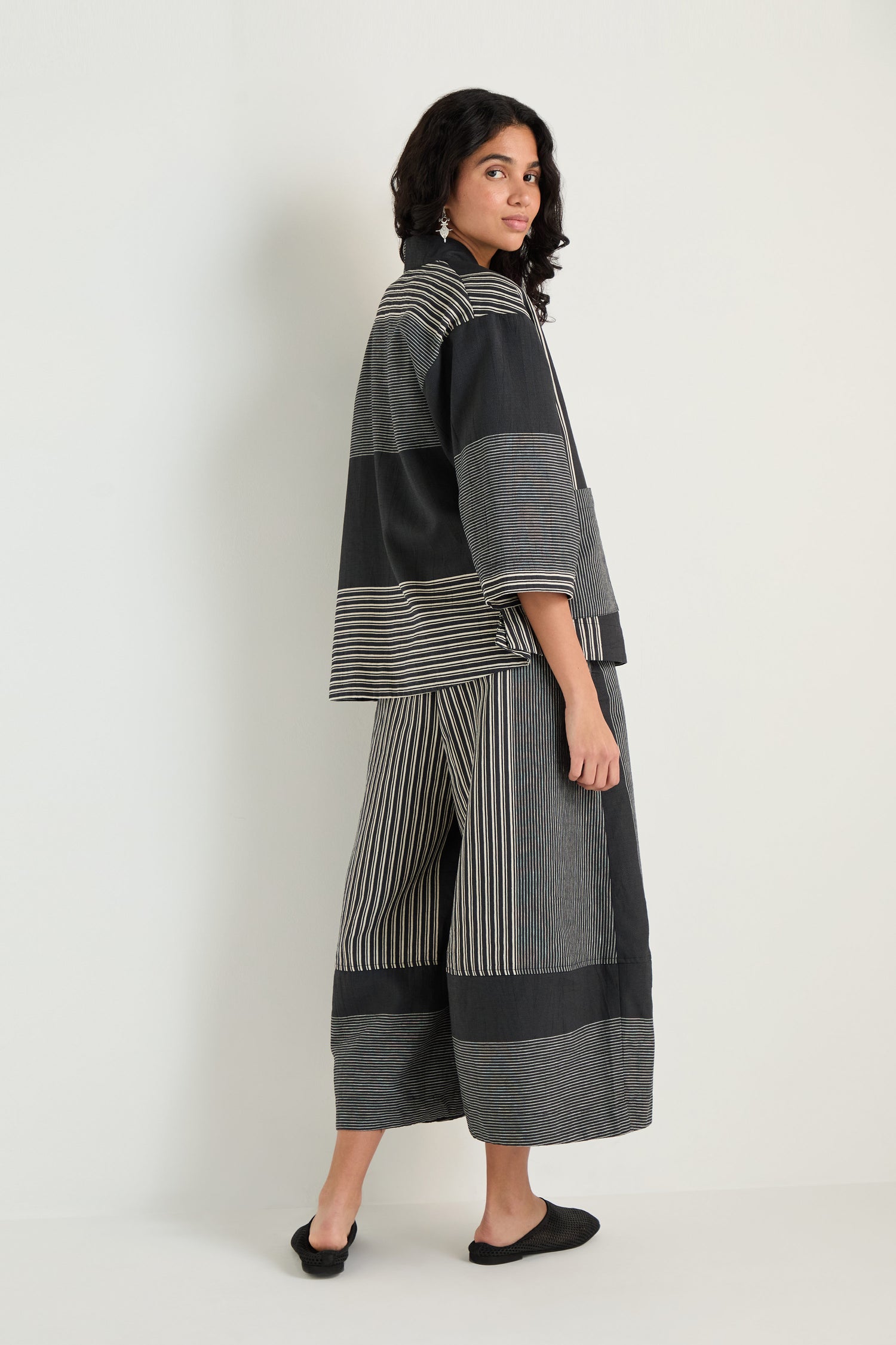 A person stands sideways against a plain light background, wearing the Mixed Stripe Easy Jacket in black and gray with wide-leg pants and black slip-on shoes.