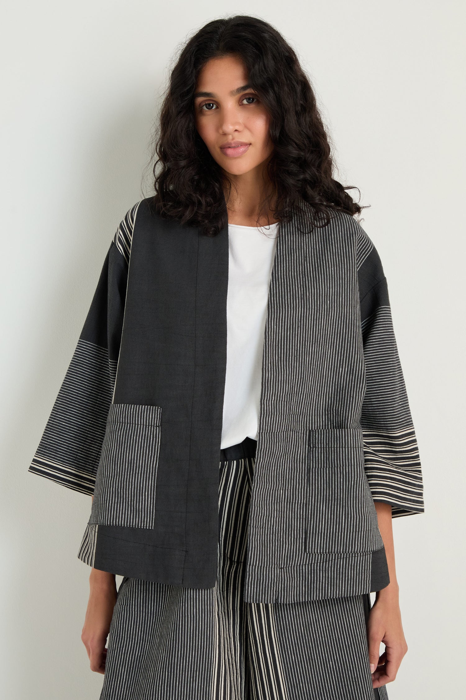 A woman stands against a plain background wearing the Mixed Stripe Easy Jacket in black and gray over a white top with matching striped pants.