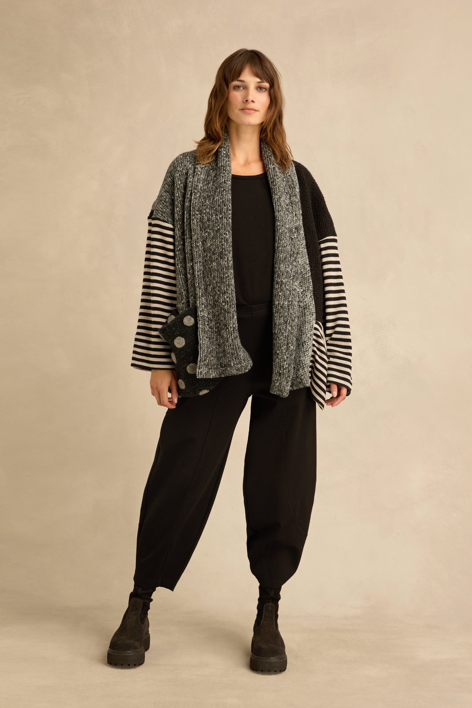 A woman stands against a beige wall wearing a black outfit, layered with the Mixed Spot Stripe Cardigan, paired with a gray scarf and black boots.