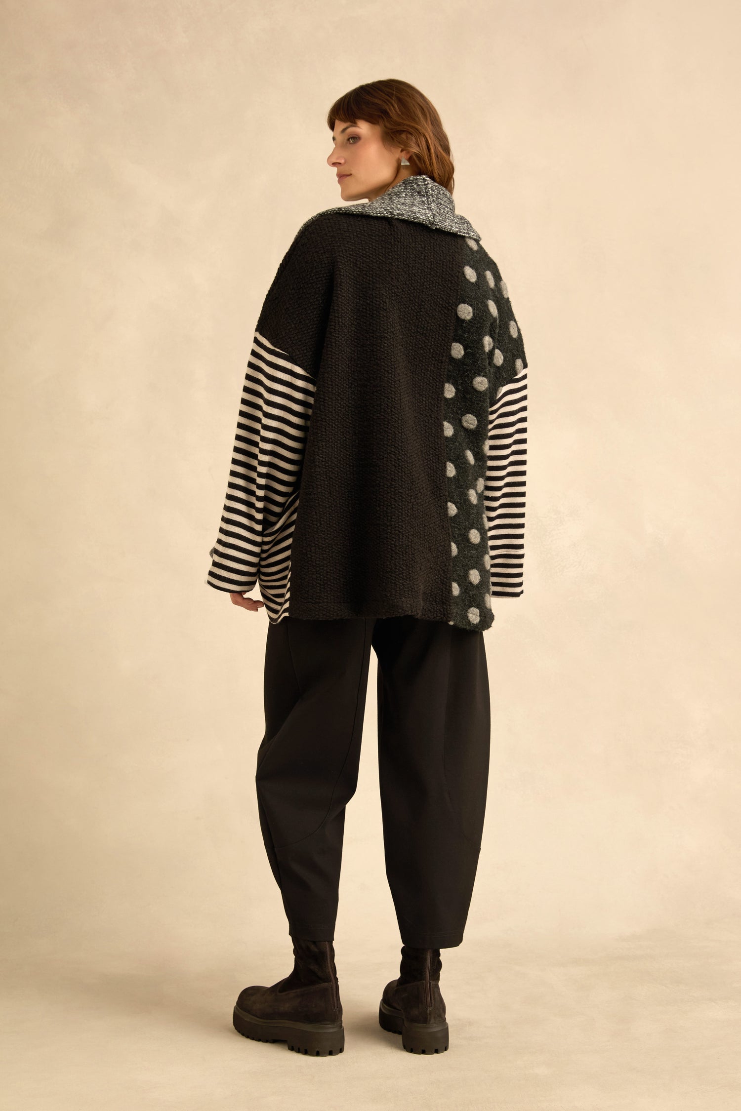 A person stands with their back to the camera, wearing the Mixed Spot Stripe Cardigan—a black and gray textured knit featuring stripes and polka dots—paired with black pants and black boots against a neutral background.