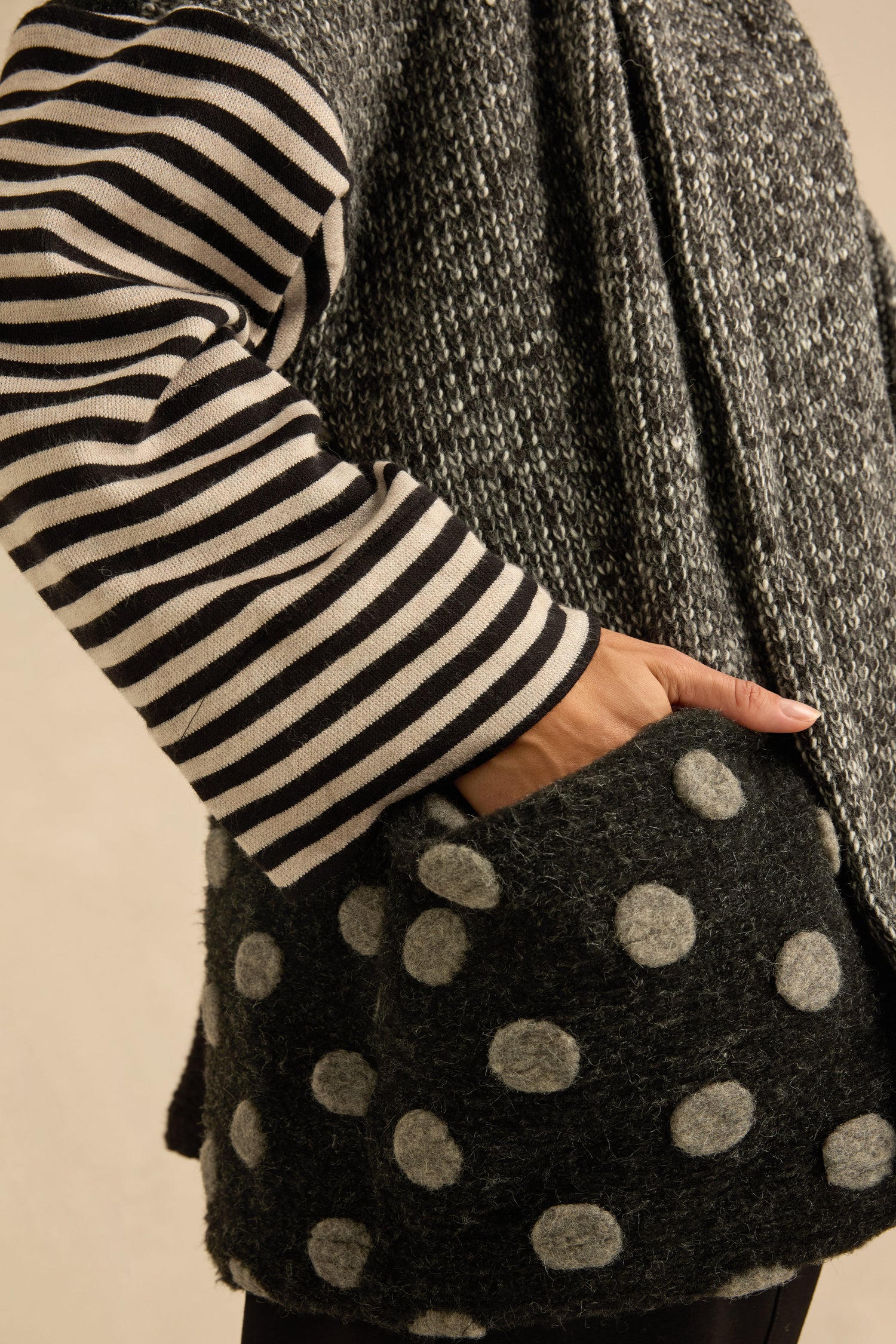 A person wears the Mixed Spot Stripe Cardigan—a textured gray piece styled with patchwork elements, featuring a large polka dot pocket and a black-and-white striped sleeve—with their hand placed inside the pocket.