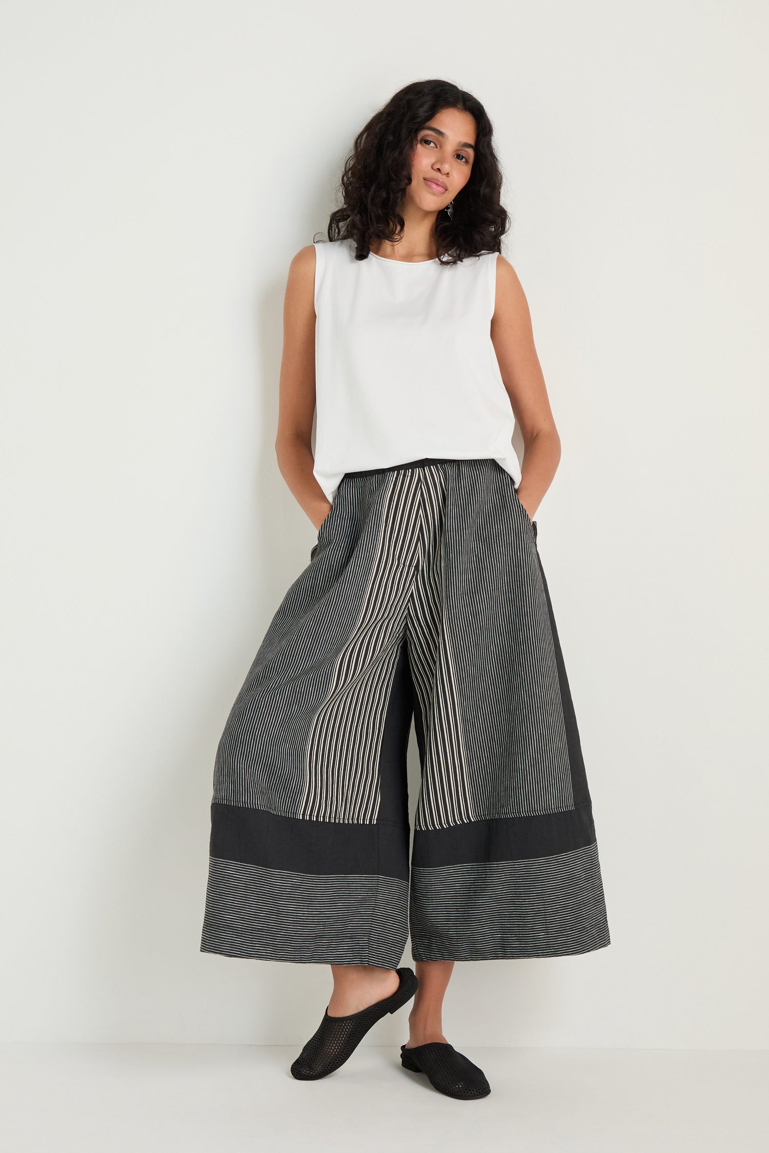 A person stands against a white wall wearing Mixed Stripe Wide Leg Trousers with an elasticated waist, a sleeveless white top, and black shoes.