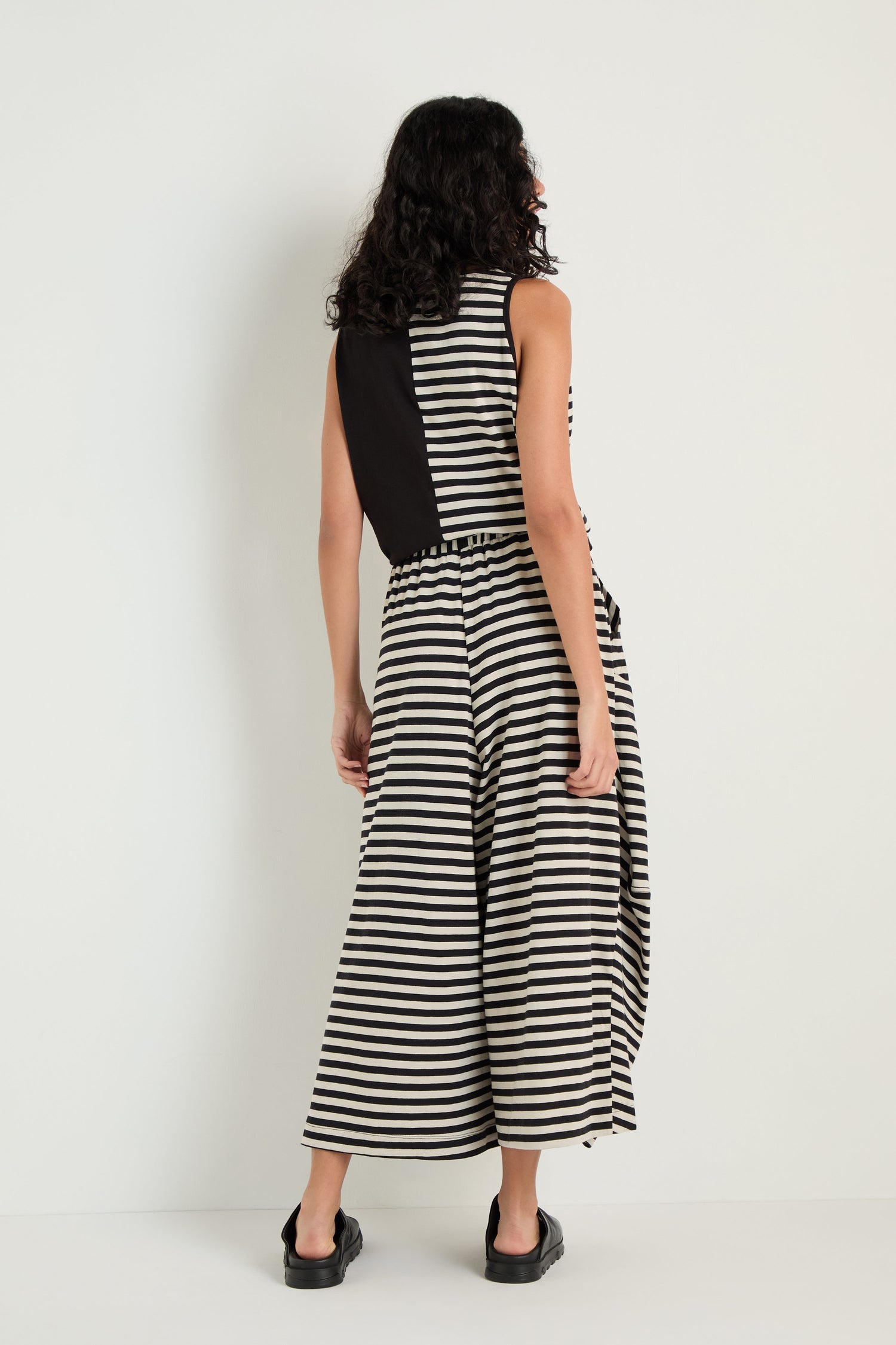 A person faces away, wearing Stripe Balloon Trousers with black shoes and a sleeveless black and white striped dress featuring a solid black back panel, creating a chic layered look.