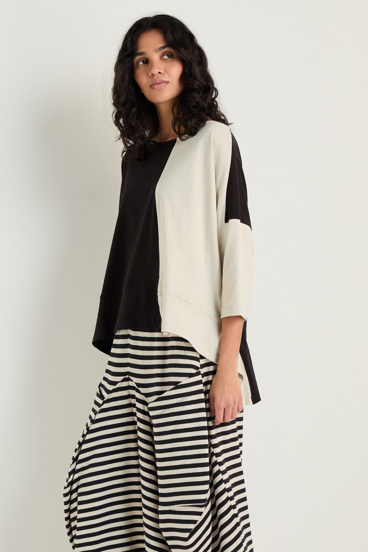 A woman stands against a plain light background wearing the Two-Tone Jersey Top with relaxed drape and three-quarter sleeves, paired with wide black-and-white striped pants.