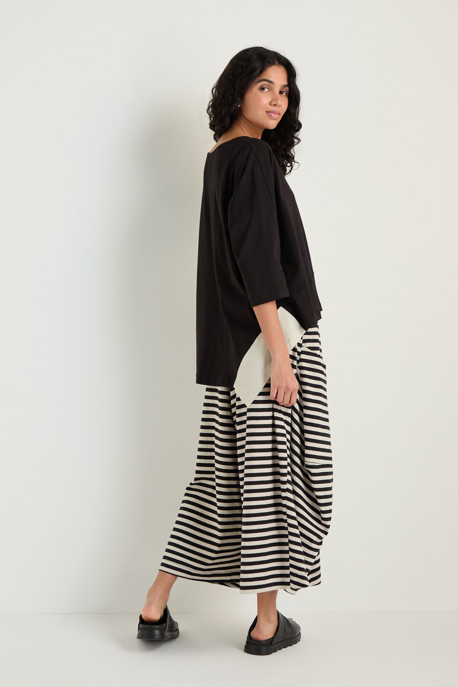 Woman stands against a plain background, wearing a relaxed black and white striped maxi skirt, black shoes, and the Two-Tone Jersey Top, looking over her shoulder.