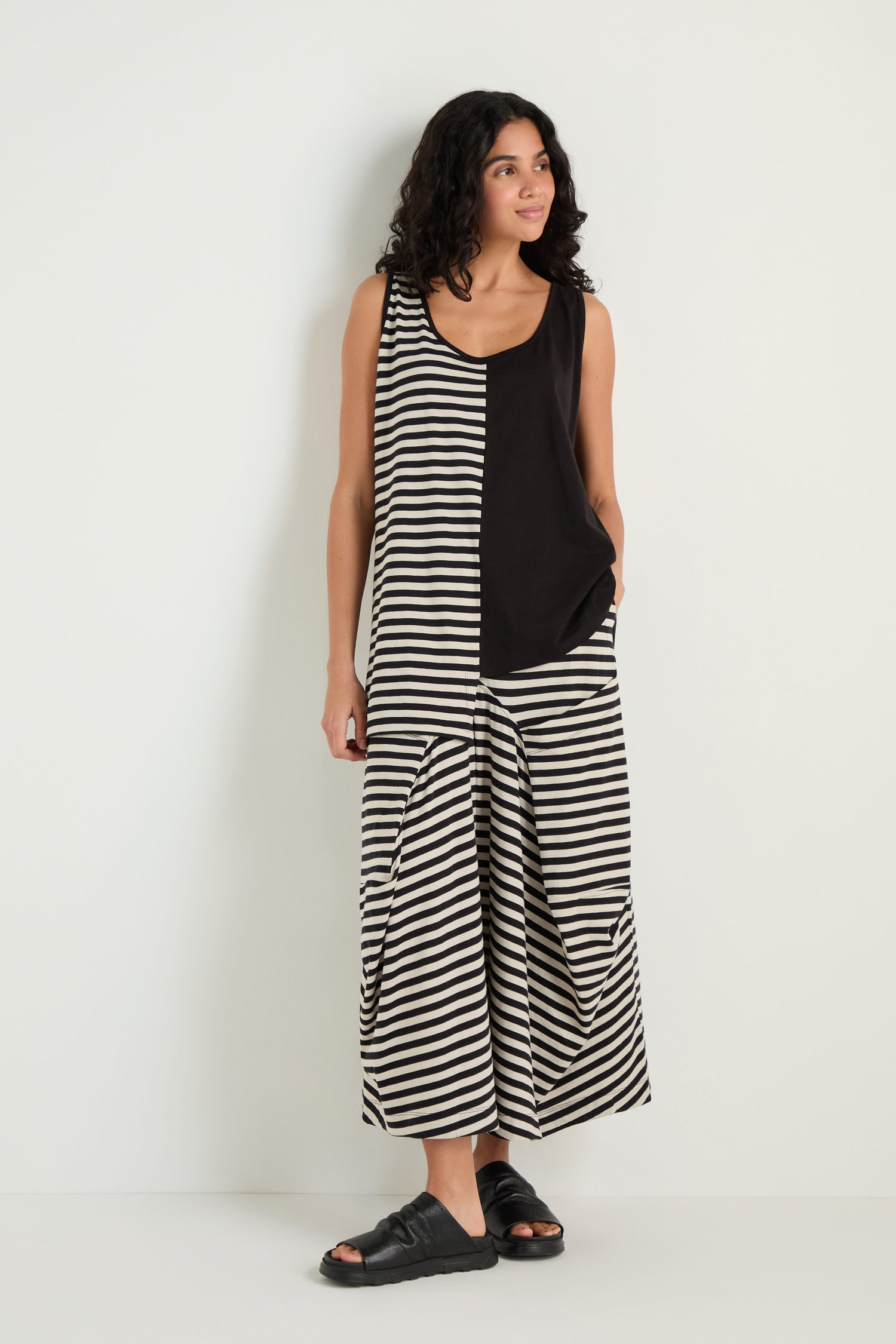 A woman stands against a white wall wearing the Stripe and Plain Sleeveless Top with wide stripe panel pants and black sandals.