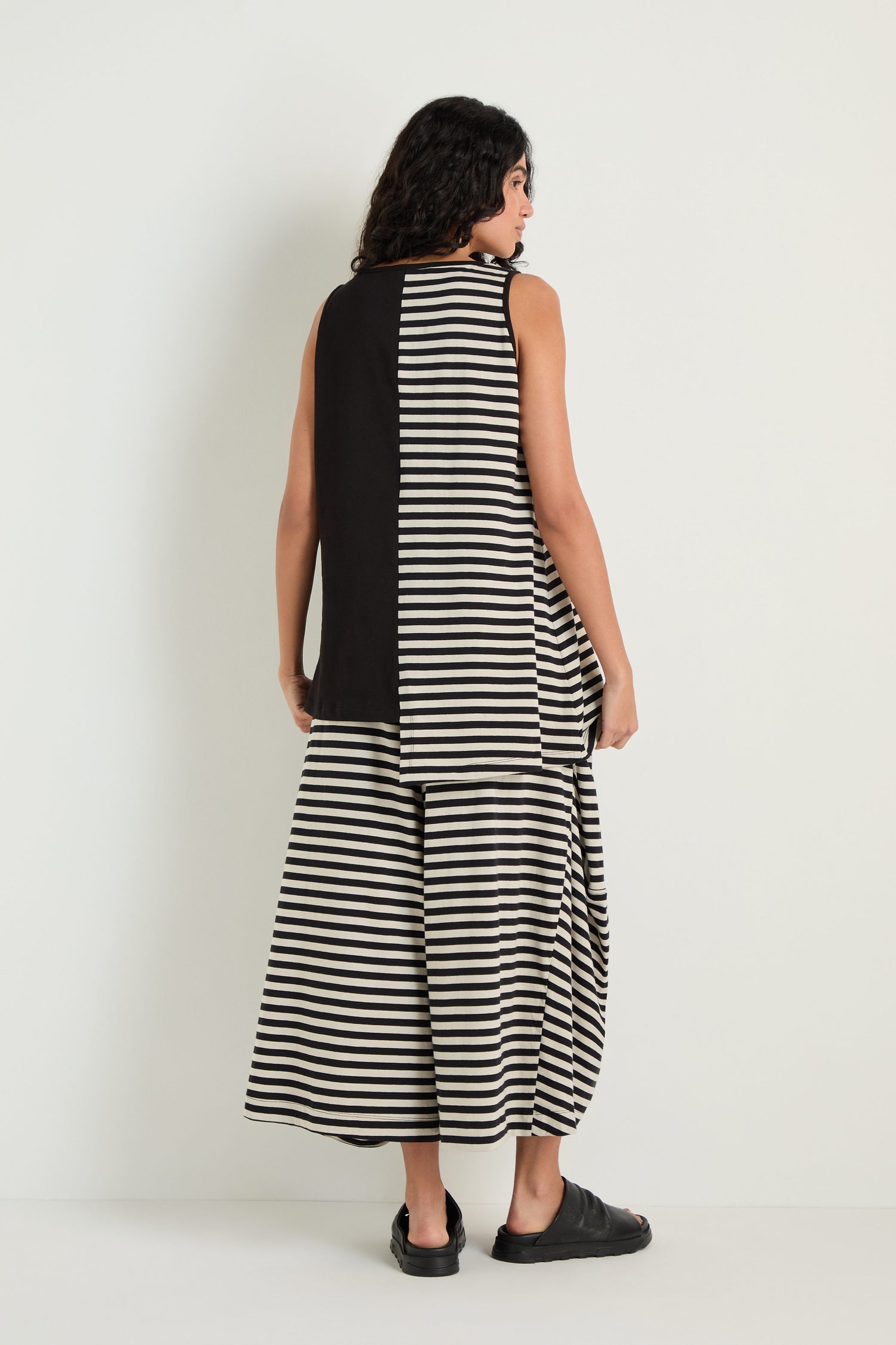 A woman stands facing away, wearing the Stripe and Plain Sleeveless Top—pure cotton, sleeveless, with a solid black panel and black-and-white stripes—paired with matching wide striped pants and black shoes.