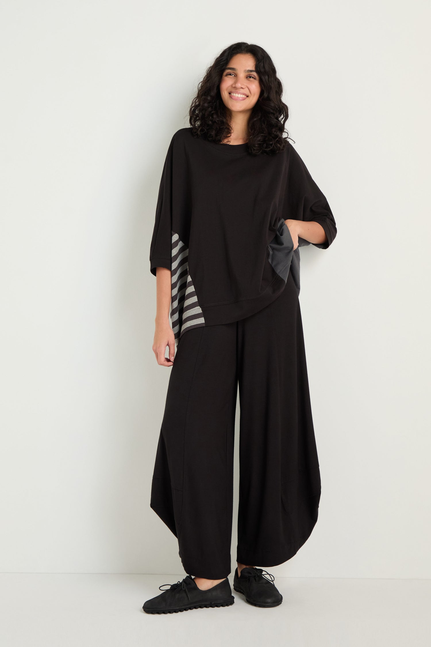 A person stands against a plain wall, wearing a Boxy Side Detail Top with striped accents and wide black pants, paired with black sneakers.