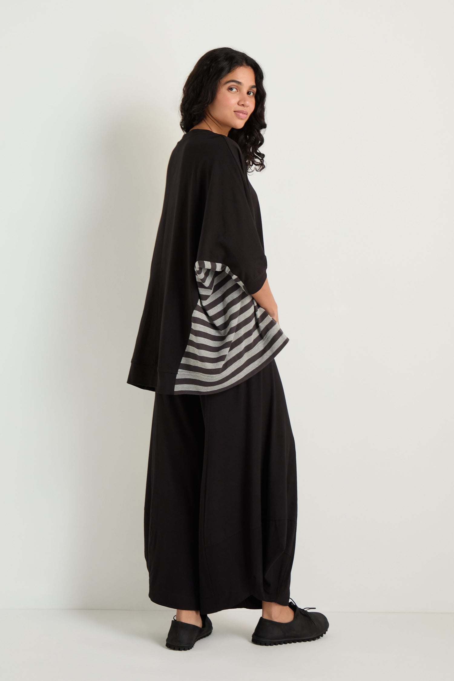 A woman stands sideways against a white wall, wearing black wide pants and the Boxy Side Detail Top with gray and black stripes, paired with black shoes.
