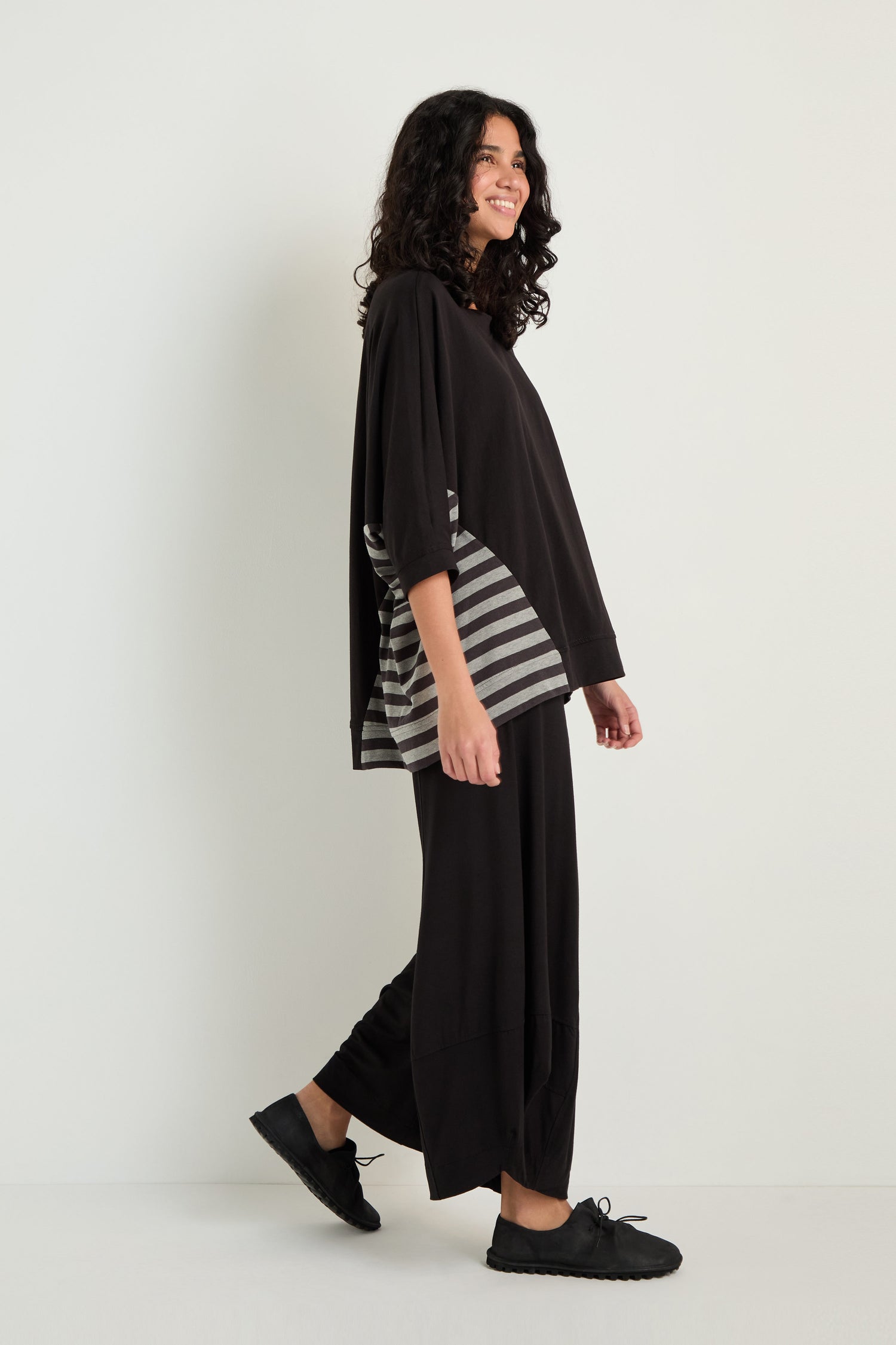 A person stands sideways, smiling, wearing the Boxy Side Detail Top with gray side stripes, wide black pants, and black shoes against a plain white background.