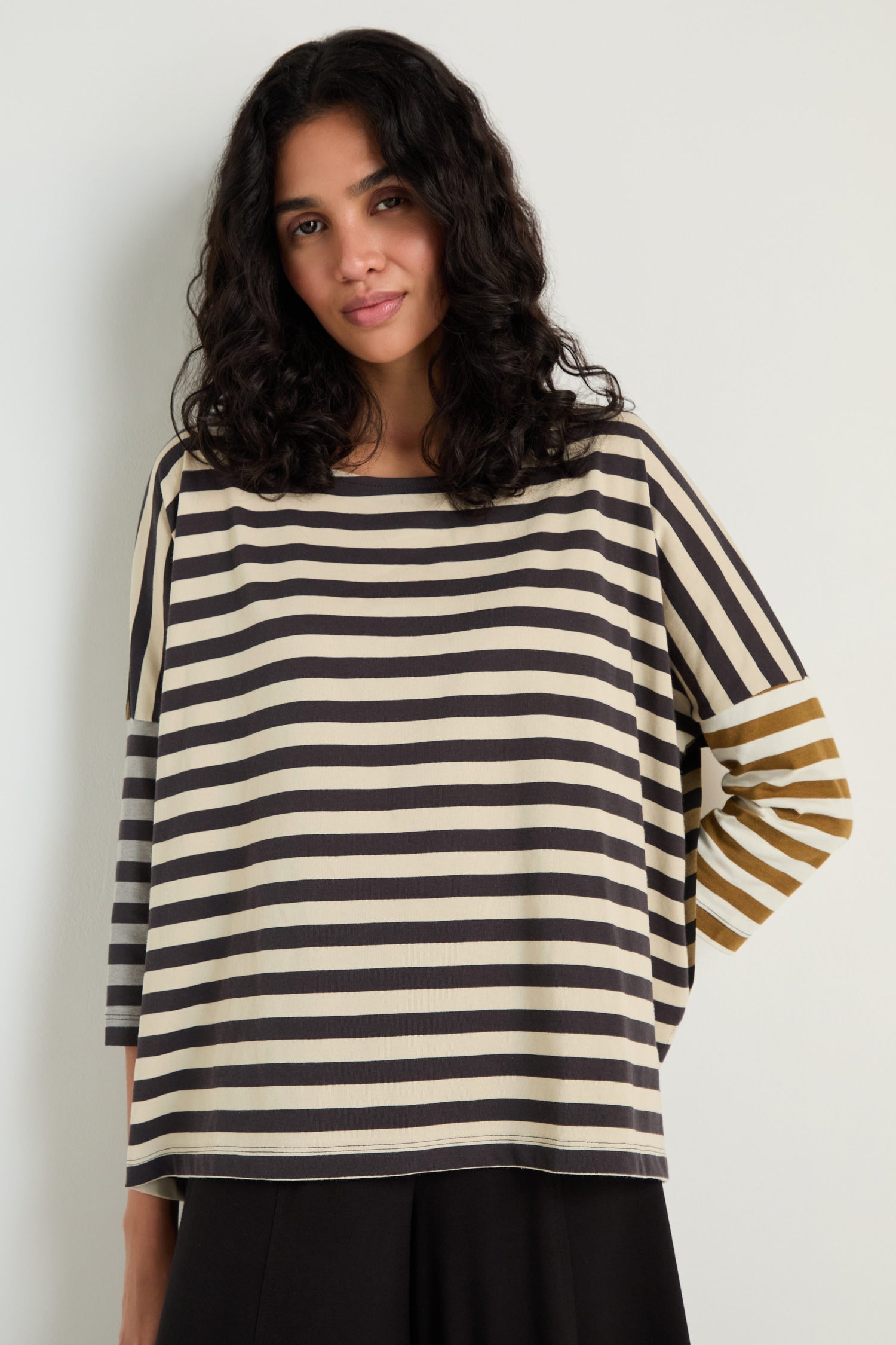 A woman with shoulder-length curly hair wears the Boxy Stripe Top in black, white, and brown tones with a relaxed silhouette and long sleeves, standing against a plain light-colored wall.