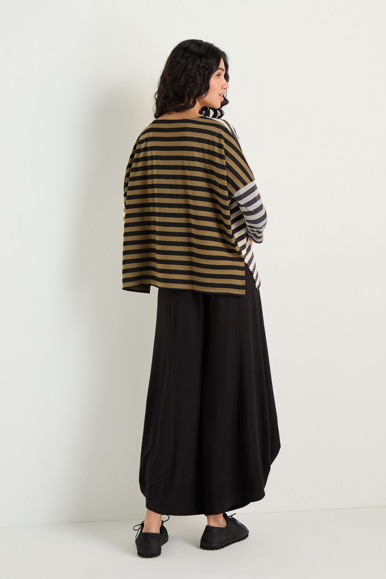 A woman with dark curly hair stands facing away, wearing the Boxy Stripe Top in olive and black, paired with a loose black skirt and black shoes for a relaxed silhouette against a plain background.