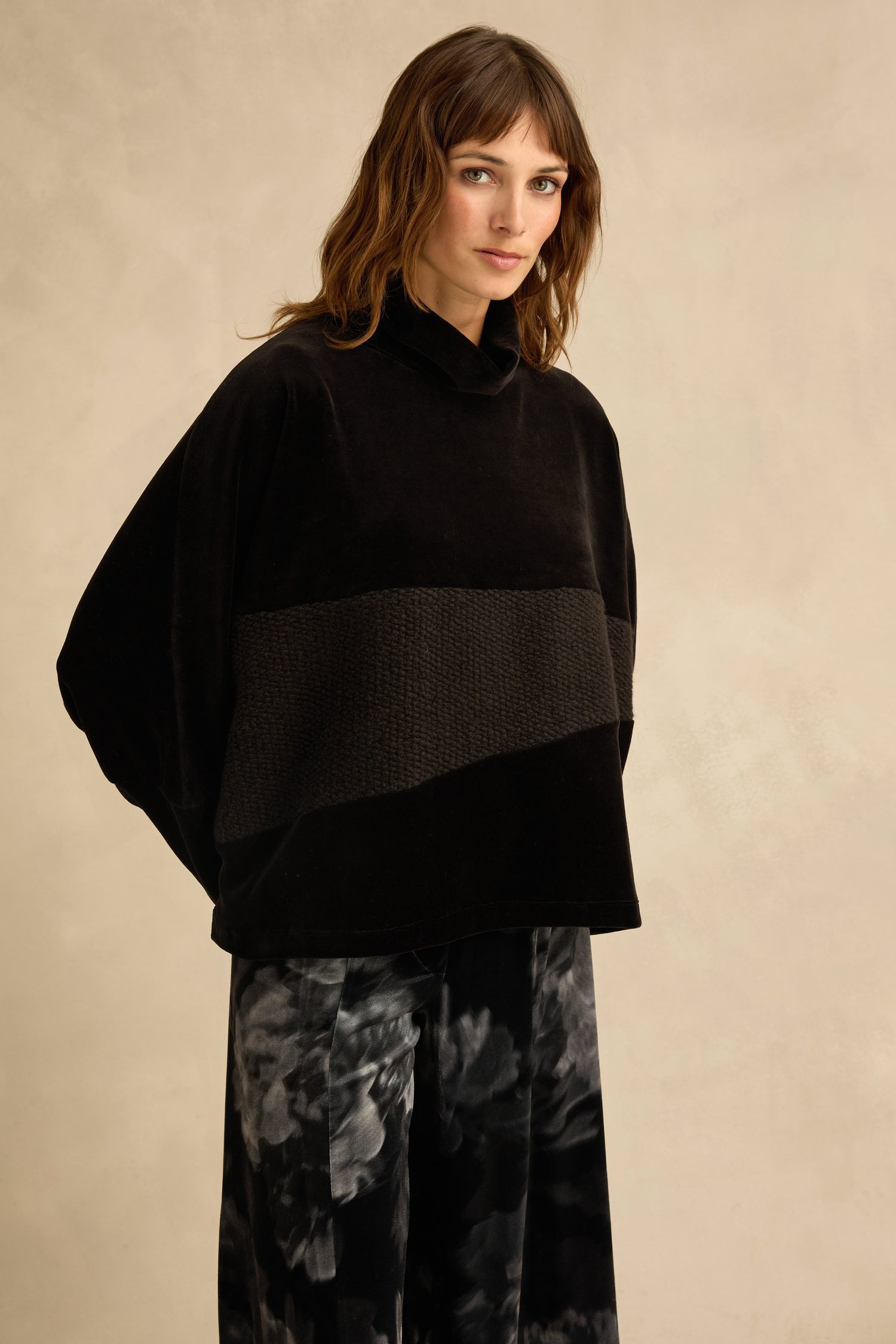 A woman wearing a Cowl Neck Velvet Jersey Top by Urban Alembika, featuring a loose black velvet jersey with a textured band, and black-and-gray patterned pants stands against a plain beige background.
