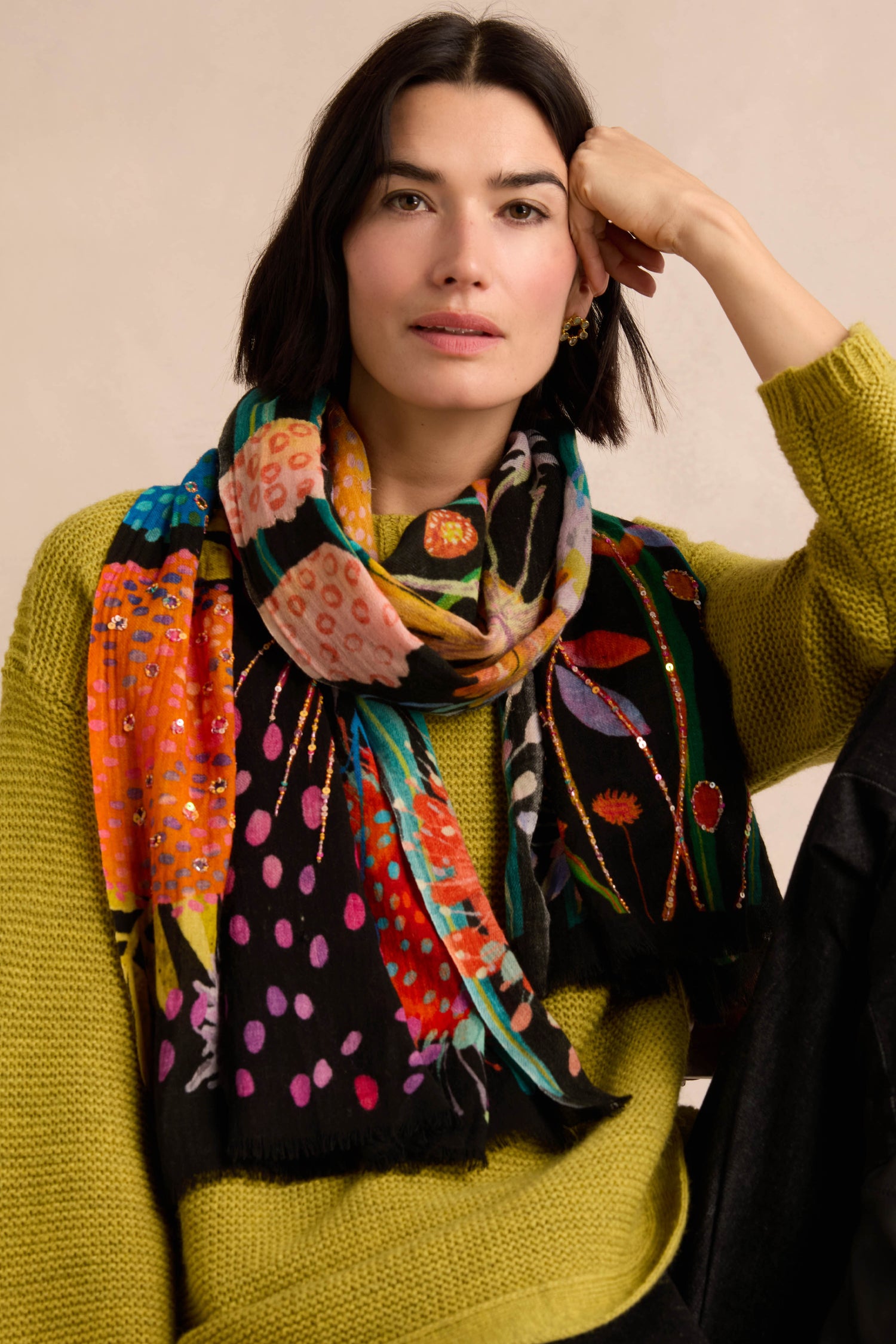 A woman with shoulder-length dark hair wears a lime green sweater and the Hand Embellished Wild Garden Wool Scarf, which features a colorful botanical print, as she sits with her arm resting on a chair.