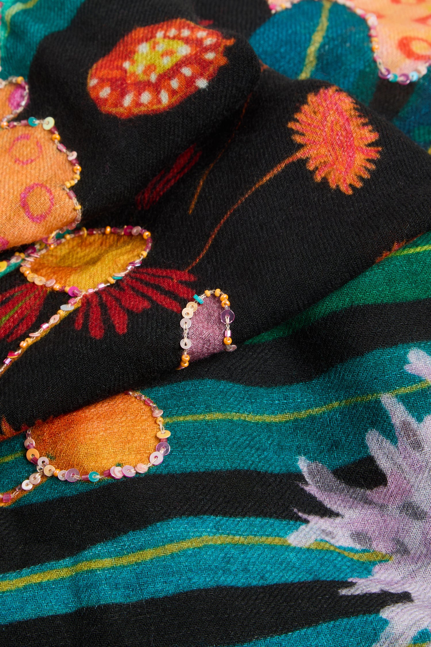 A close-up of the Hand Embellished Wild Garden Wool Scarf showcases black wool adorned with a vibrant botanical print, featuring colorful floral patterns, green stripes, and small sequins accentuating some of the flowers.