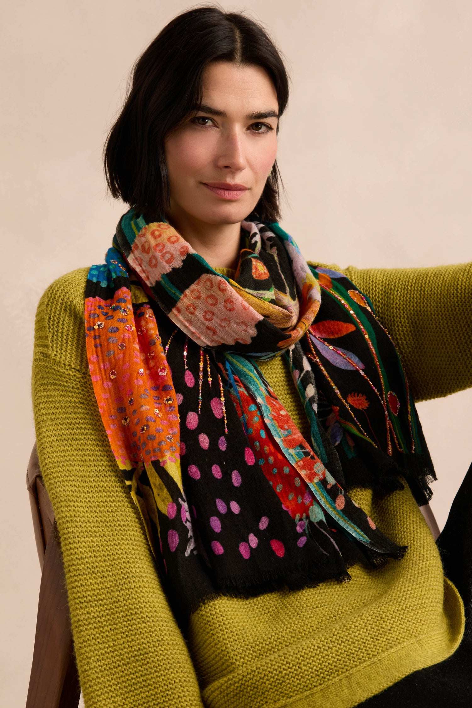 A person with dark hair wears a green knit sweater and the Hand Embellished Wild Garden Wool Scarf while sitting against a neutral background.