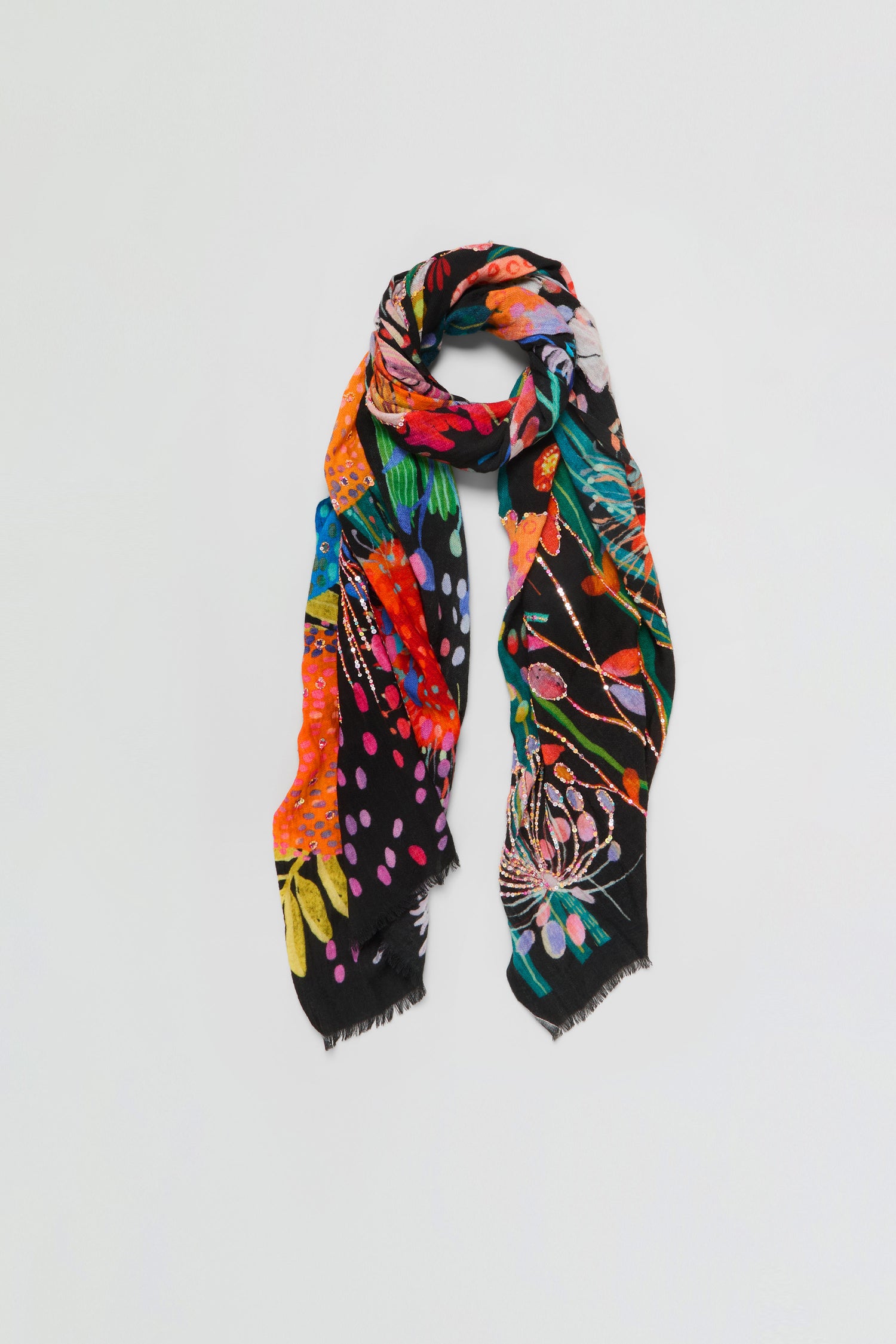 The Hand Embellished Wild Garden Wool Scarf, featuring a black base with a multicolored floral and abstract pattern and finished with frayed edges, is loosely draped and displayed against a plain white background.