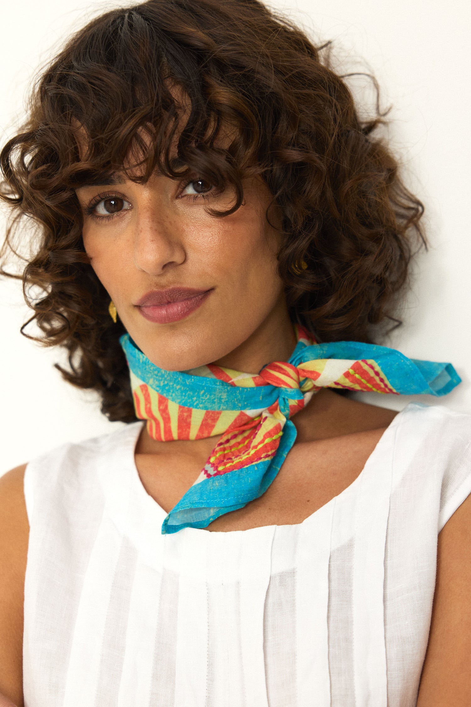 A woman with curly brown hair wears a white sleeveless top and the Embroidered Parasols Cotton Bandana, featuring a colorful blue and red print, tied around her neck as she stands against a plain background.