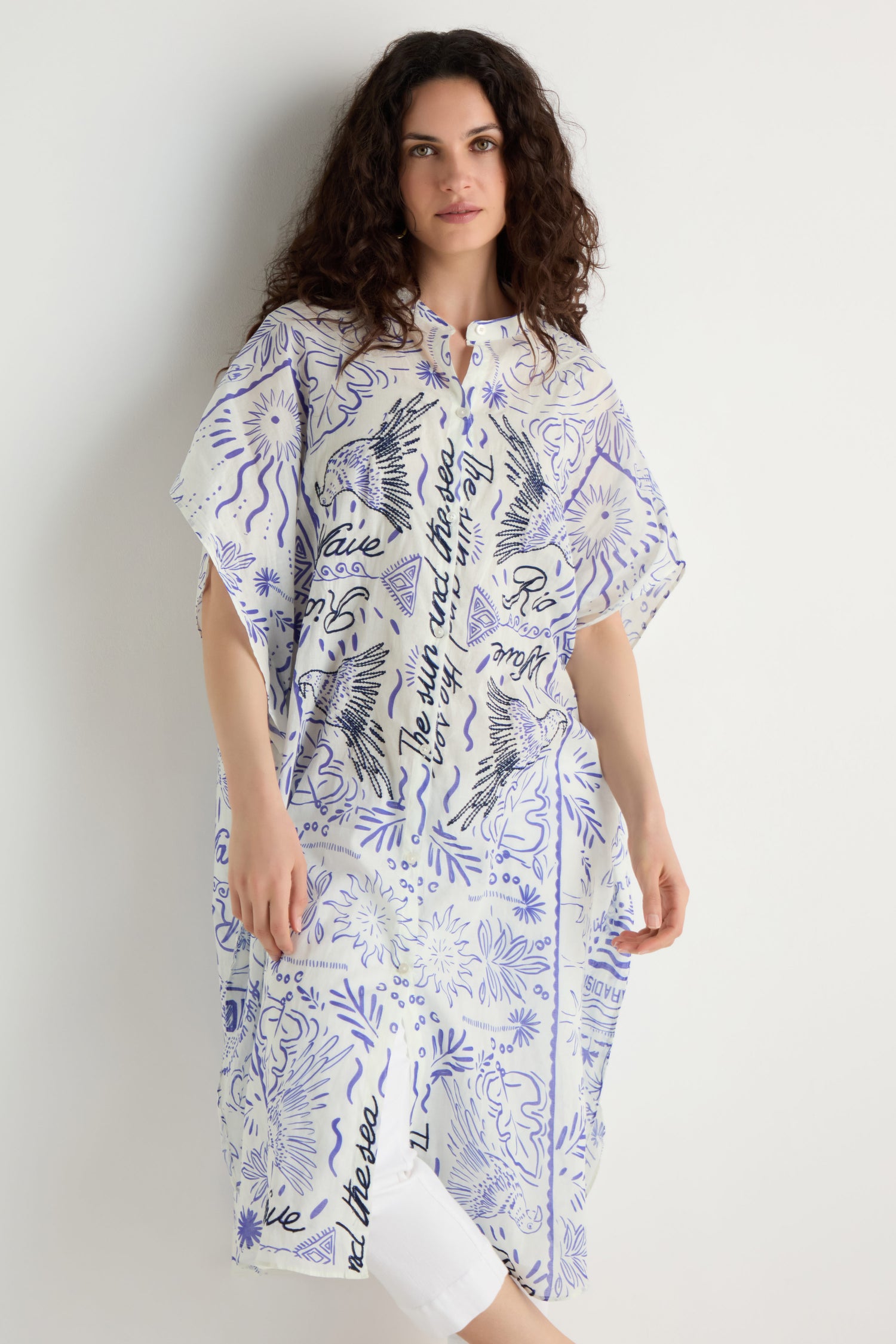 A woman stands against a white background wearing the Paradiso Cotton Shirt Dress, a loose white and blue patterned kaftan-style dress with text, floral designs, and embroidered detailing.