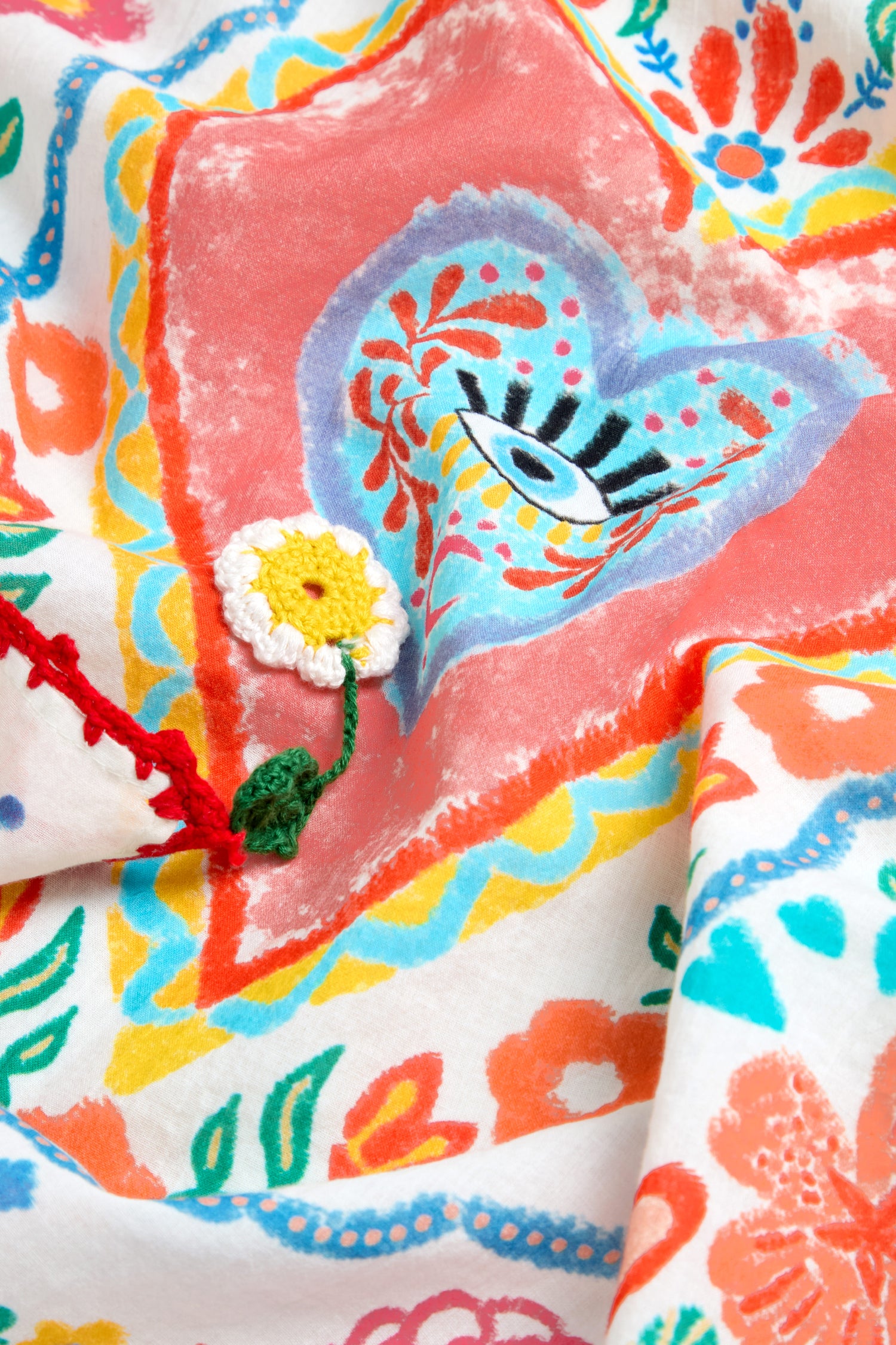 A close-up of the Embroidered Love In Bloom Cotton Bandana, showcasing vibrant floral patterns, a heart and eye motif, and a small crocheted yellow and white flower with a green stem.