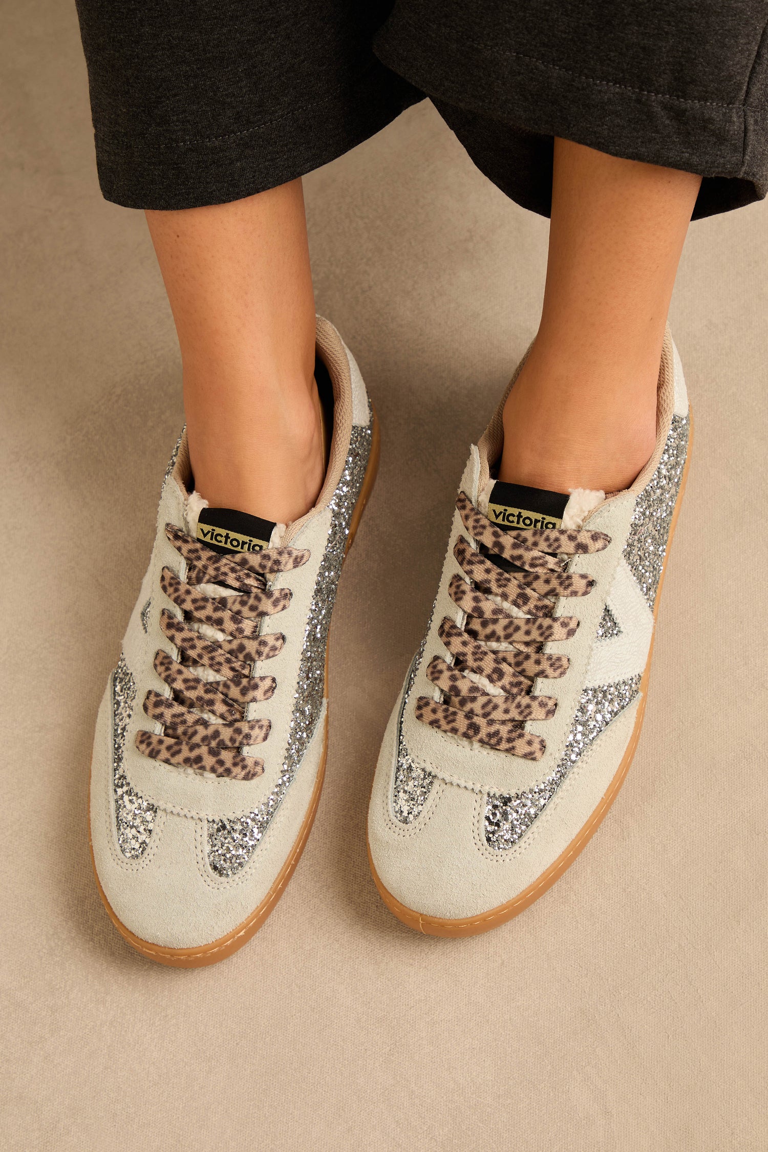 A person wearing Berlin Glitter Trainers, featuring light-colored uppers with glittery sides, animal-print heel detail, leopard print laces, suede panels, and gum soles, standing on a neutral floor.