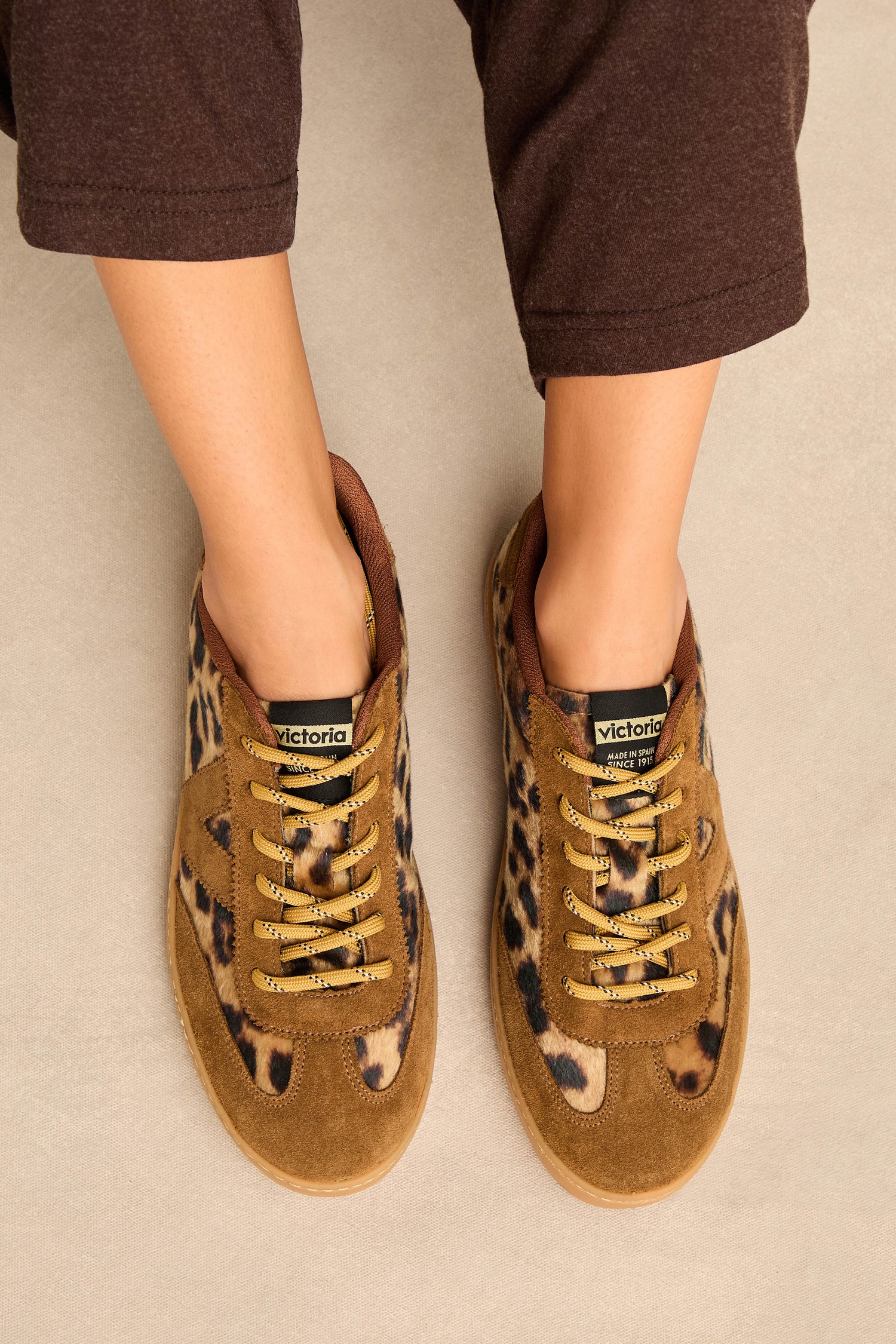 A person sits on a beige surface, viewed from above, wearing brown pants and the Berlin Animal Print Trainers with suede panel accents.