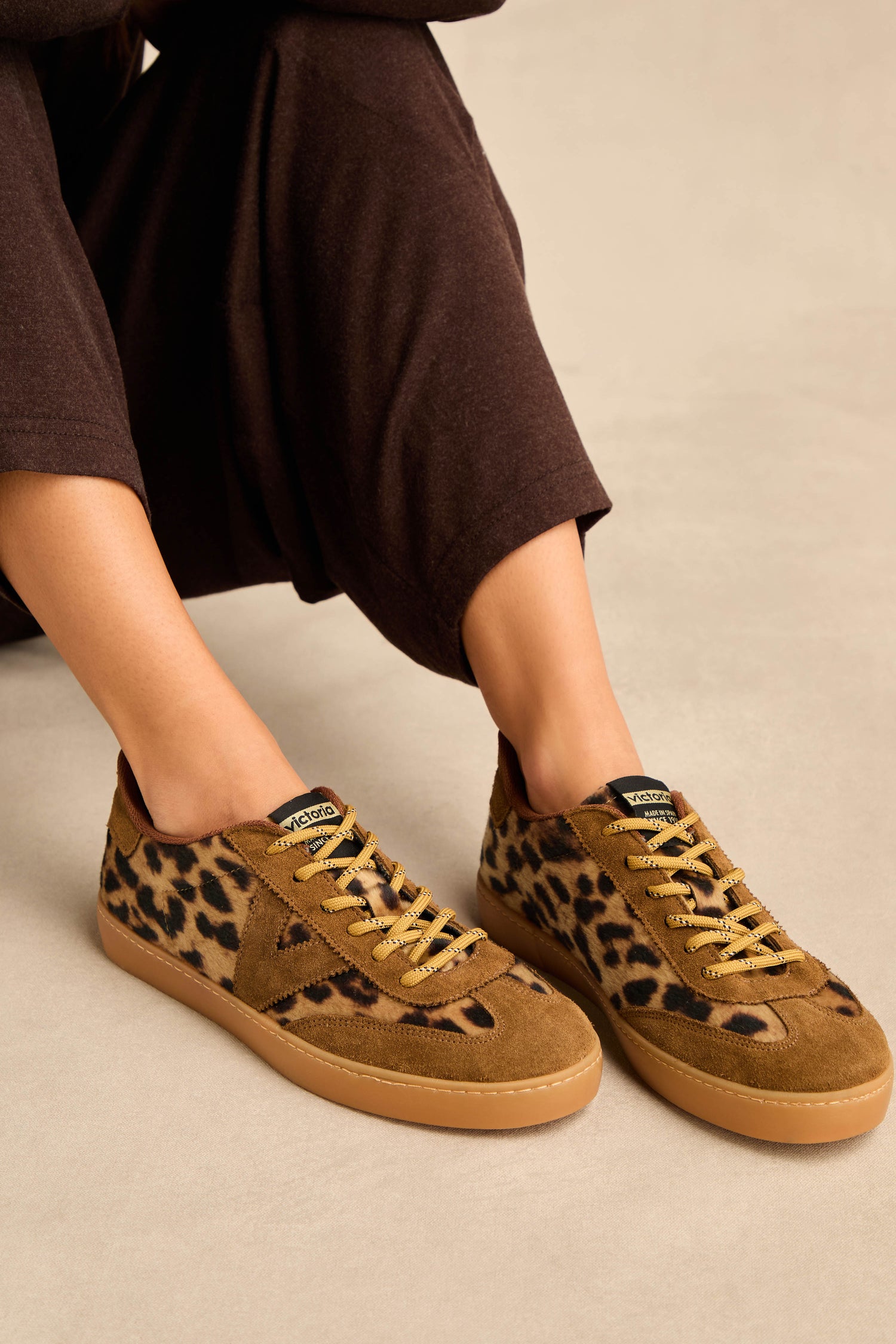 Wearing the Berlin Animal Print Trainers—featuring brown suede panels, leopard print accents, and tan laces—a person sits on a beige surface with brown pants visible. These stylish trainers are made in Spain.
