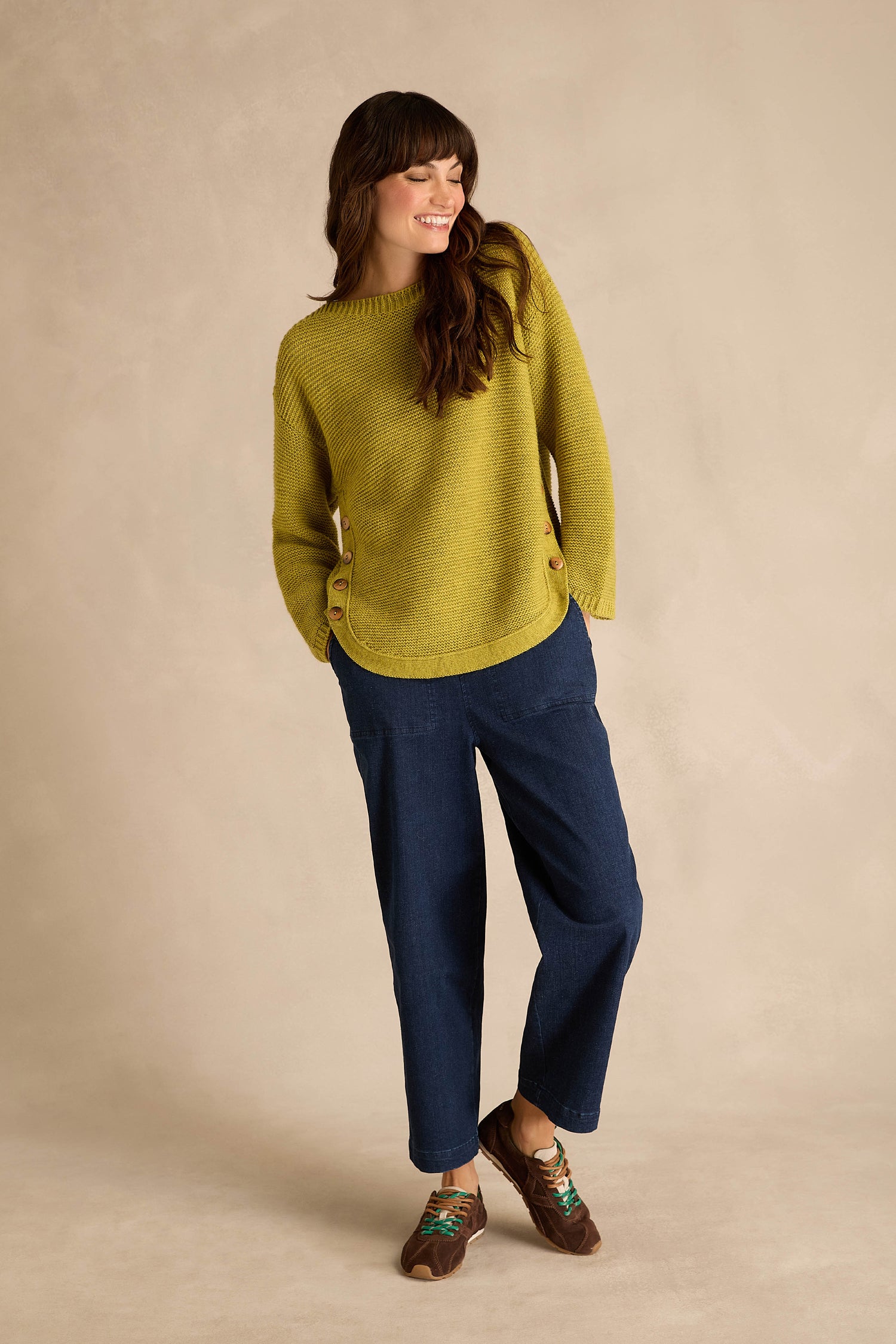A woman stands against a neutral background, smiling and looking to the side, dressed in a green sweater, blue pants, and our Aster Suede Trainers featuring contrast stitching.