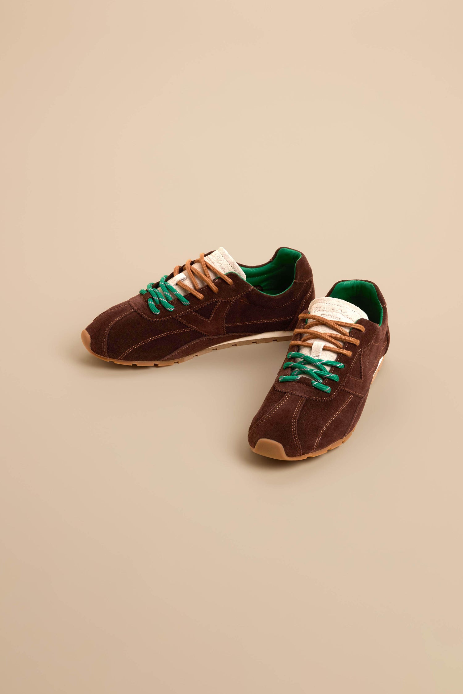 A pair of Aster Suede Trainers in brown cowhide suede featuring green inner lining, beige soles, contrast stitching, and mismatched beige and green laces, displayed on a beige background.