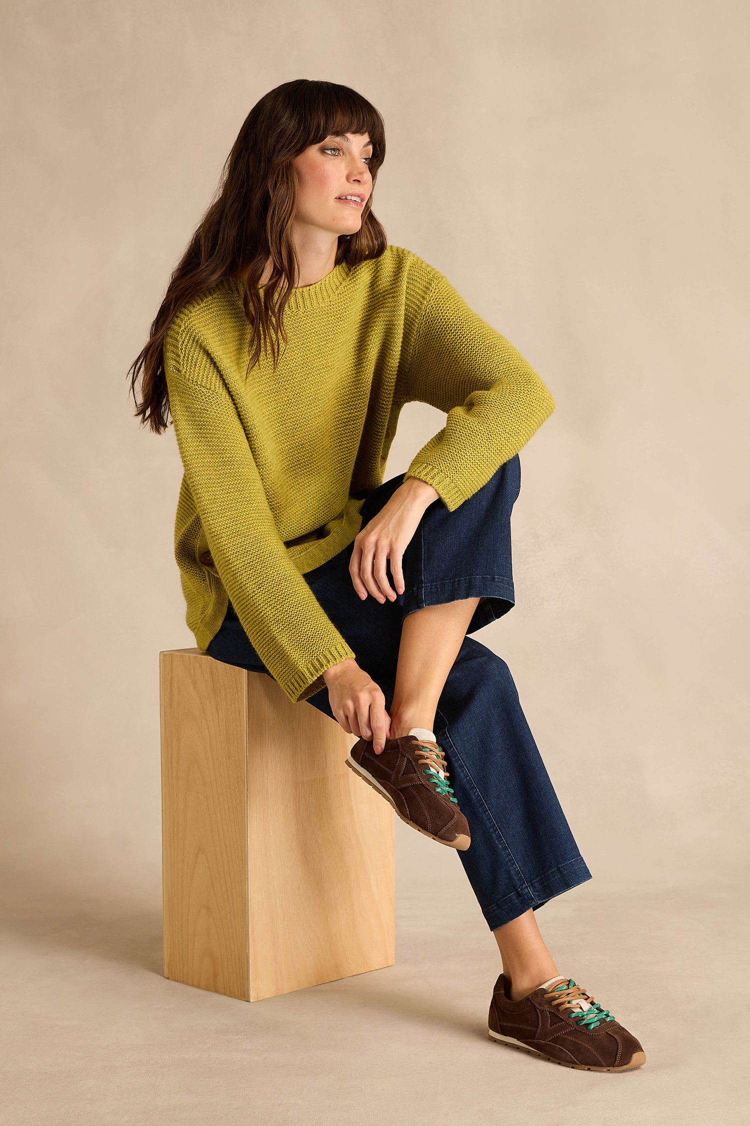 A woman sits on a wooden block against a beige background, wearing blue jeans, a yellow-green sweater, and the Aster Suede Trainers—brown suede shoes featuring multicolored laces and contrast stitching.