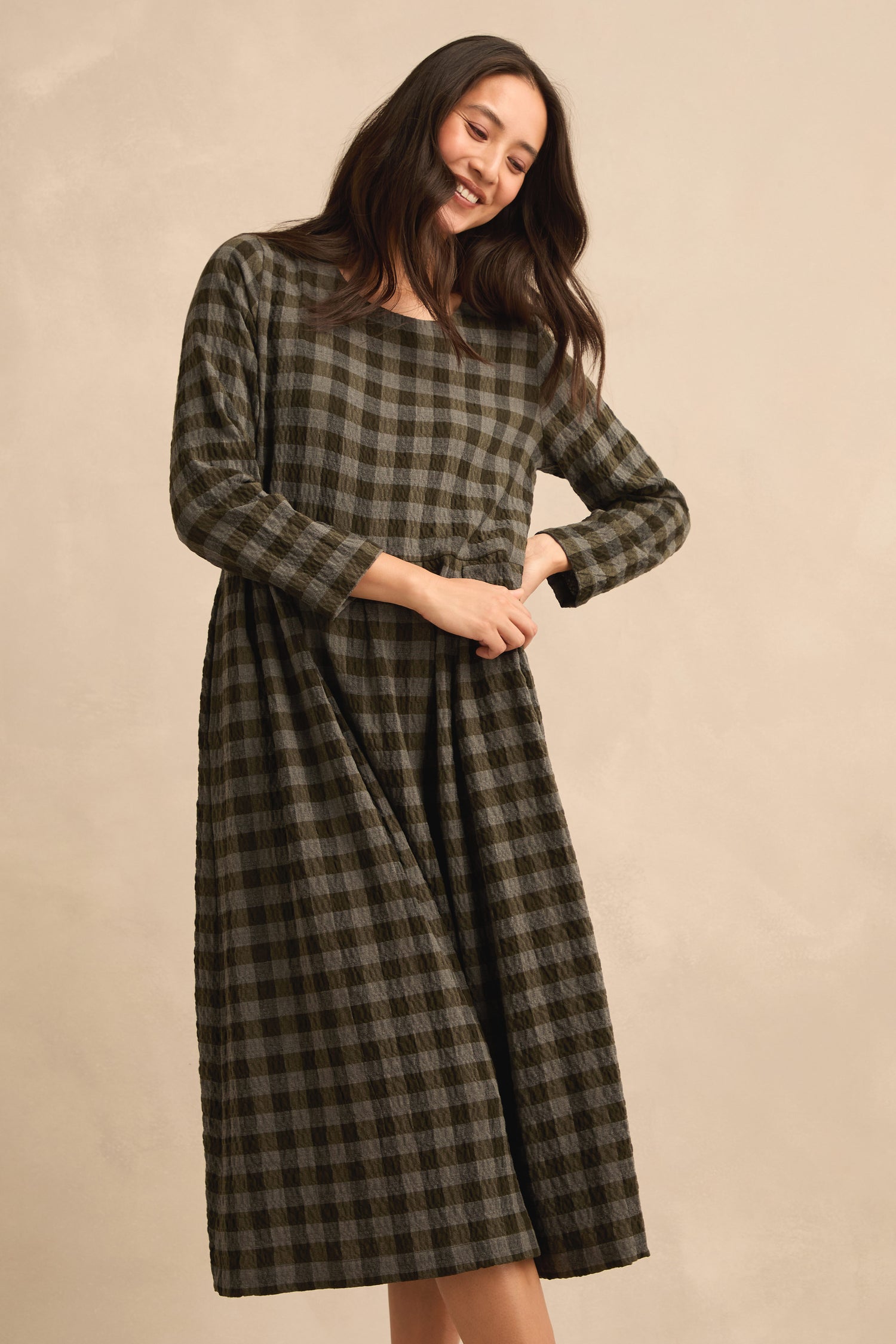 A woman stands against a beige background, smiling with her hands clasped in front, wearing the Crinkle Check Flared Dress in green and black with long sleeves.
