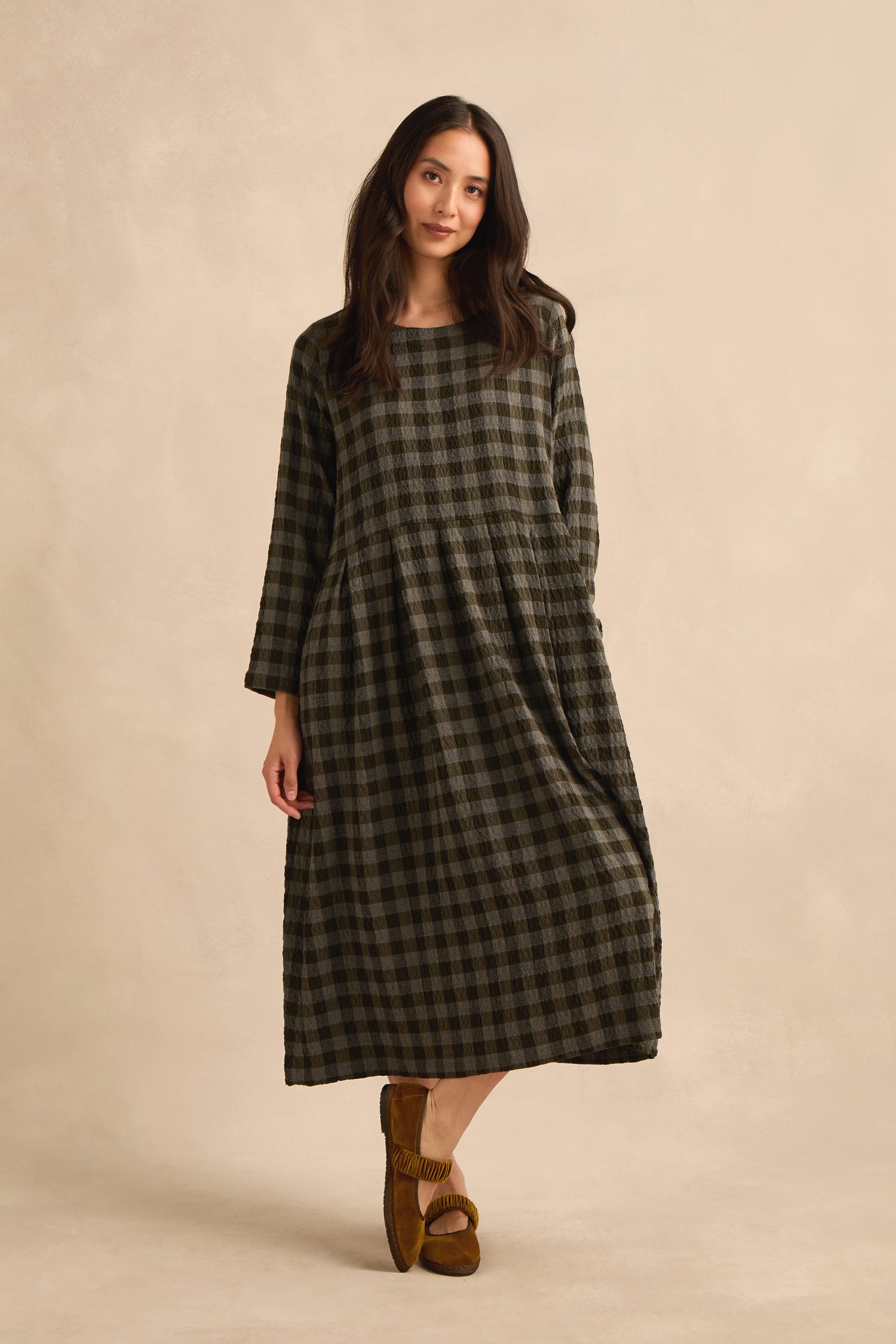 A woman stands against a neutral background wearing the Crinkle Check Flared Dress in dark tones, paired with brown shoes.