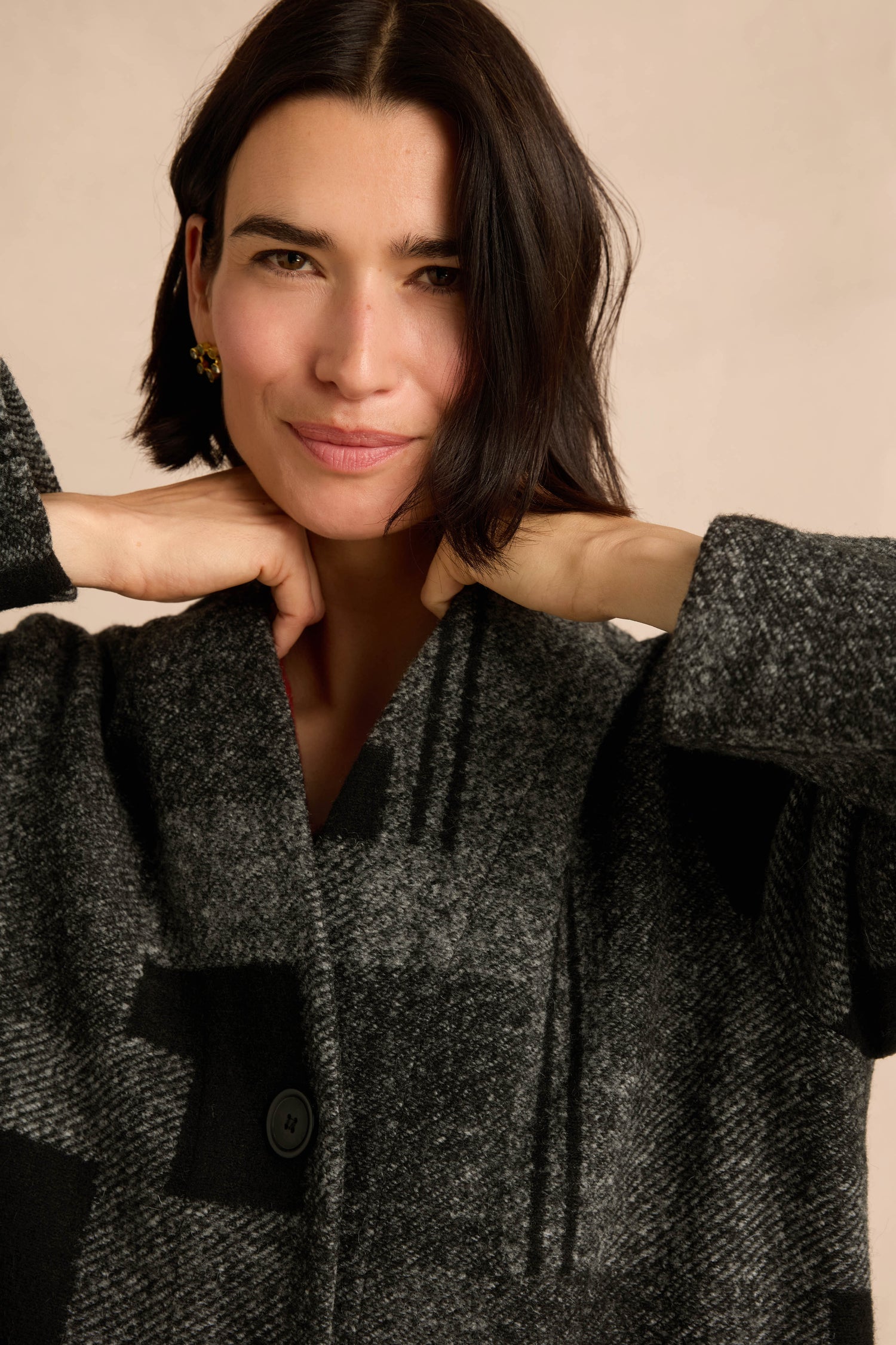 A woman with short dark hair wears the Melange Patchwork Wool Coat, gently holding the collar with both hands and smiling softly at the camera against a light beige background.