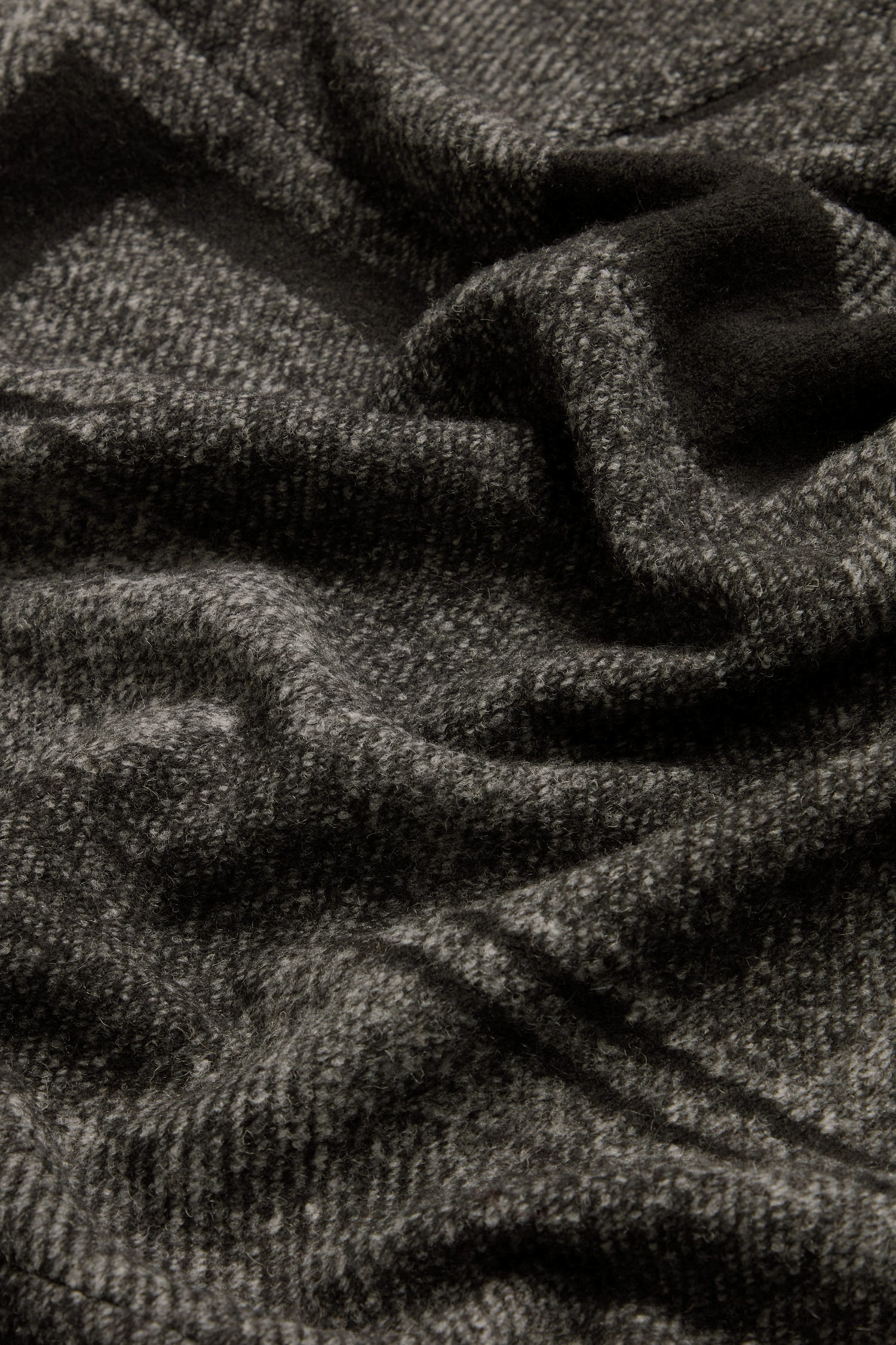 A close-up of the Melange Patchwork Wool Coat features its dark gray wool fabric, highlighting the soft, textured surface with visible folds and creases.