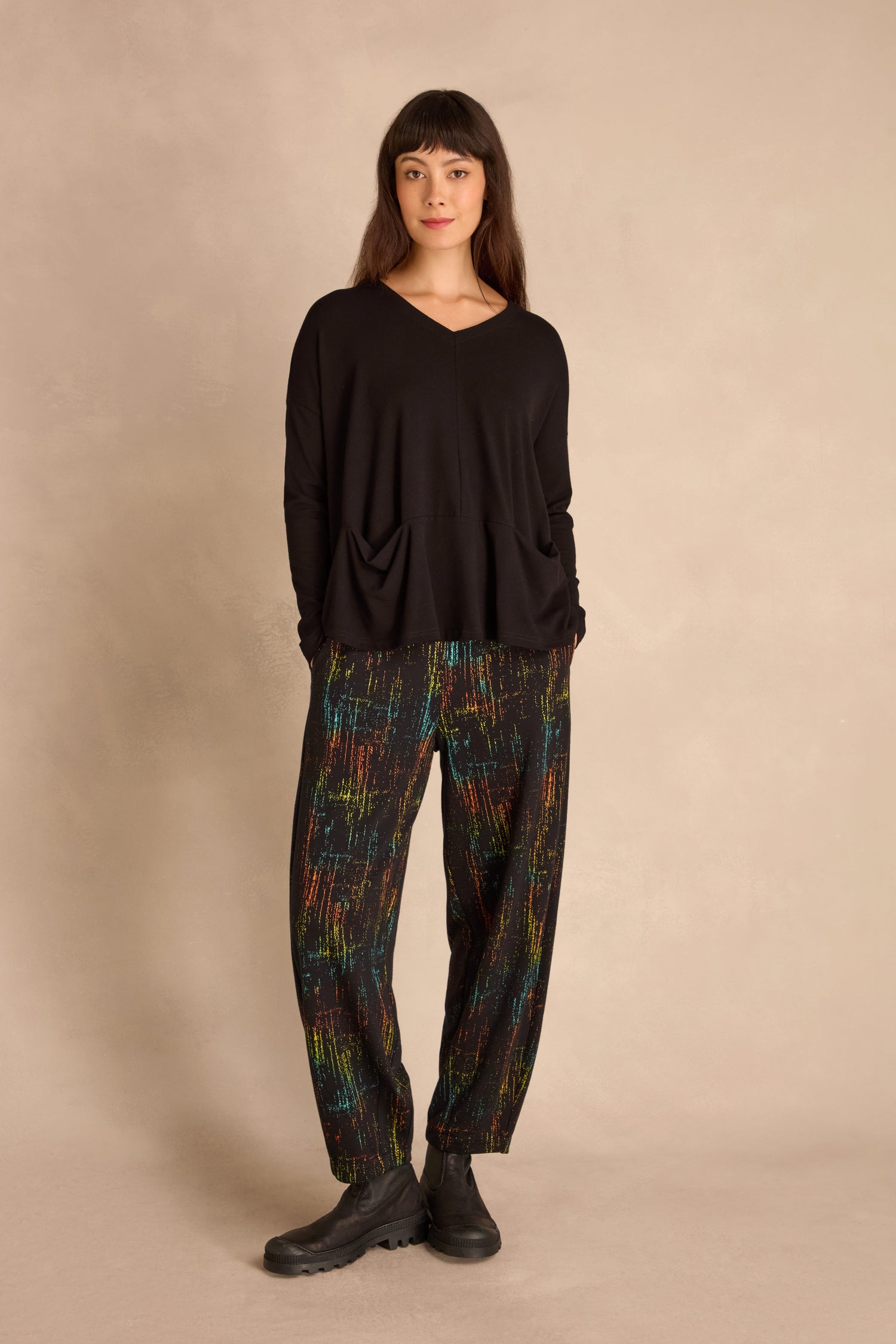 Woman standing against a beige background, wearing a loose black long-sleeve top with front pockets, Space Dye Jacquard Bubble Trousers, and black shoes.