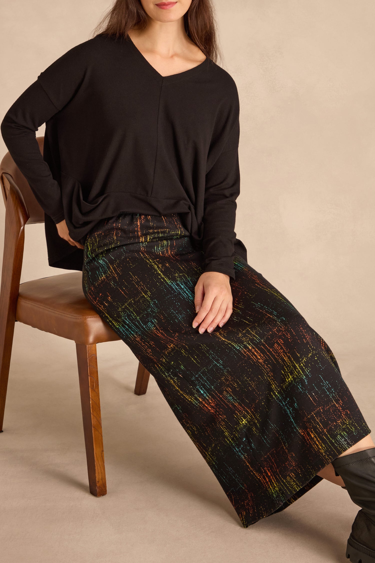 Woman seated on a wooden chair wearing a loose black long-sleeve top and the Space Dye Jacquard Tube Skirt with colorful abstract patterns.